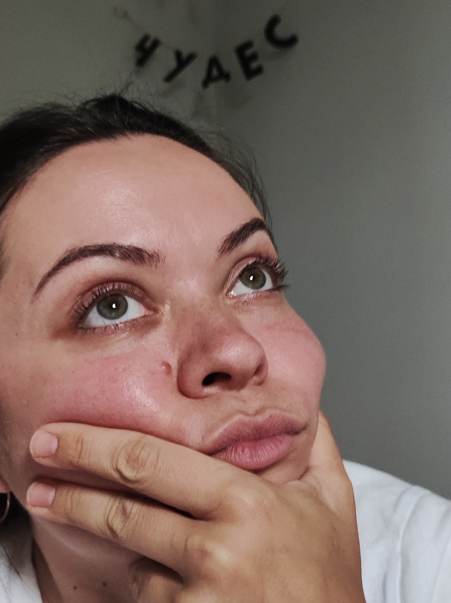 A close-up, upward-angled portrait features a woman captured in a moment of contemplation, looking upwards and to the right. Her right hand is positioned under her chin, with fingers gently resting against her cheek and mouth, enhancing her thoughtful expression. She has distinct light green eyes, complemented by dark eyelashes and well-defined eyebrows. Her lips are slightly pursed. Her skin displays some natural redness on her cheeks and around her nose, and a small mole is visible on her nose. Her dark hair is styled away from her face, and a portion of a white t-shirt is visible on her right side. A subtle hoop earring is discernible in her left ear. The background consists of a plain, solid grey wall. Affixed to the upper part of the wall are dark letters spelling out the Cyrillic word "ЧУДЕС". The lighting is soft and even, suggesting an indoor setting. The overall atmosphere conveys introspection and wonder.