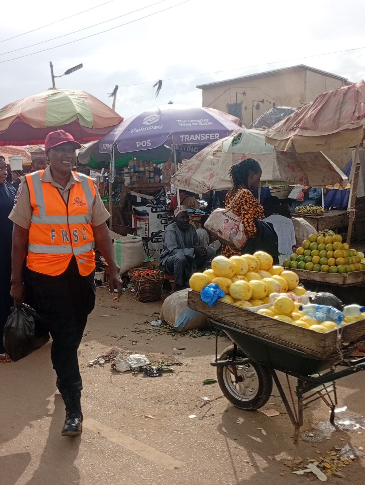 A person wearing a red cap and an orange FRSC vest over a tan shirt walks from left to right in a market. The person is carrying a black plastic bag in their left hand and is wearing black trousers and black boots. Behind the person, several stalls are visible with umbrellas providing shade. One stall features a purple umbrella with "PalmPay POS TRANSFER" written on it. A wheelbarrow filled with yellow fruits, possibly melons, is positioned in the foreground to the right of the person. Behind the wheelbarrow, a tiered display of green fruits, likely limes or lemons, is visible. Other market-goers are seen in the background, including a man seated in a gray garment and another person wearing a patterned dress and carrying a bag. The ground appears to be paved with dirt and some debris. The scene is set outdoors in what is described as the city of Rafin Par, Nigeria.