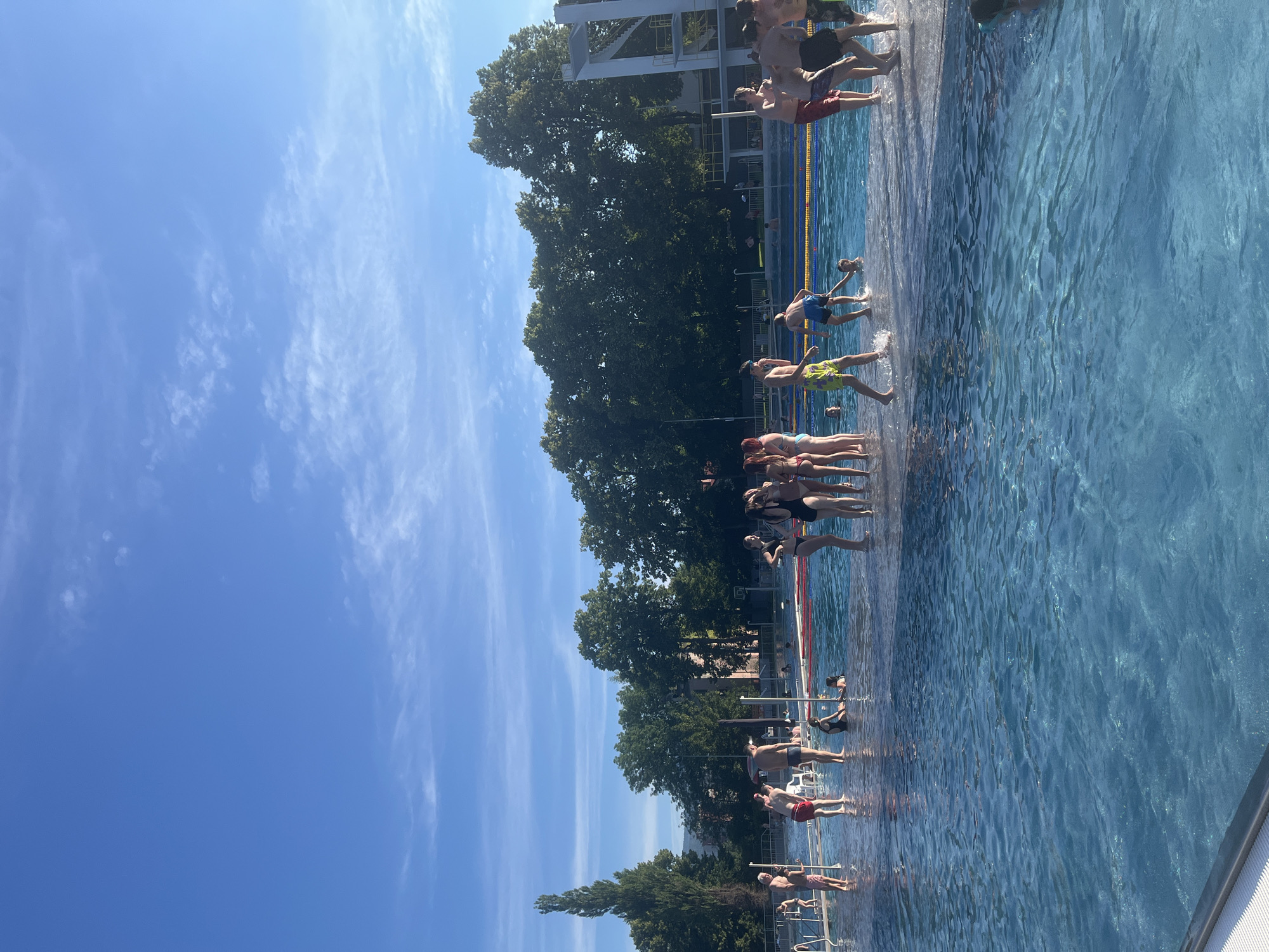 A group of people are gathered on the edge of a bright blue swimming pool under a clear blue sky with scattered white clouds. The sun is shining, casting reflections on the water. Some individuals are standing on a raised platform surrounding the pool, while others are partially submerged in the water. Trees with lush green foliage line the background, and a tall, slender tree stands prominently on the left side of the image. A diving platform structure is visible in the distance on the right. The scene appears to be a public swimming pool or a water park on a sunny day.