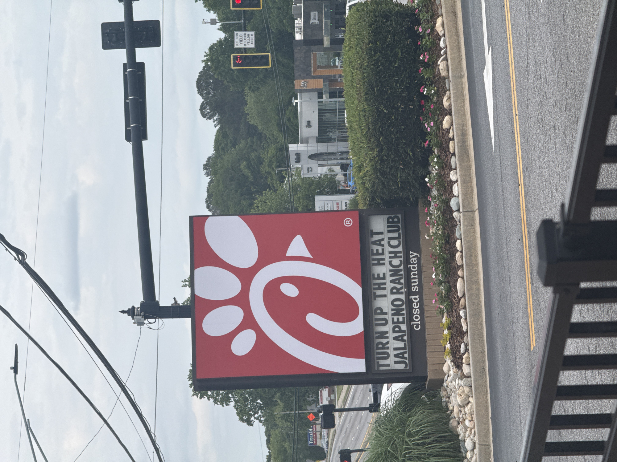 This image shows a Chick-fil-A sign in Stone Mountain, GA. The sign advertises a Jalapeno Ranch Club, and indicates that the restaurant is closed on Sunday. In the background, one can see traffic signals, trees, and other commercial buildings. The image was likely taken from an elevated position. The street is paved with double yellow lines, a white directional arrow, and a curb.