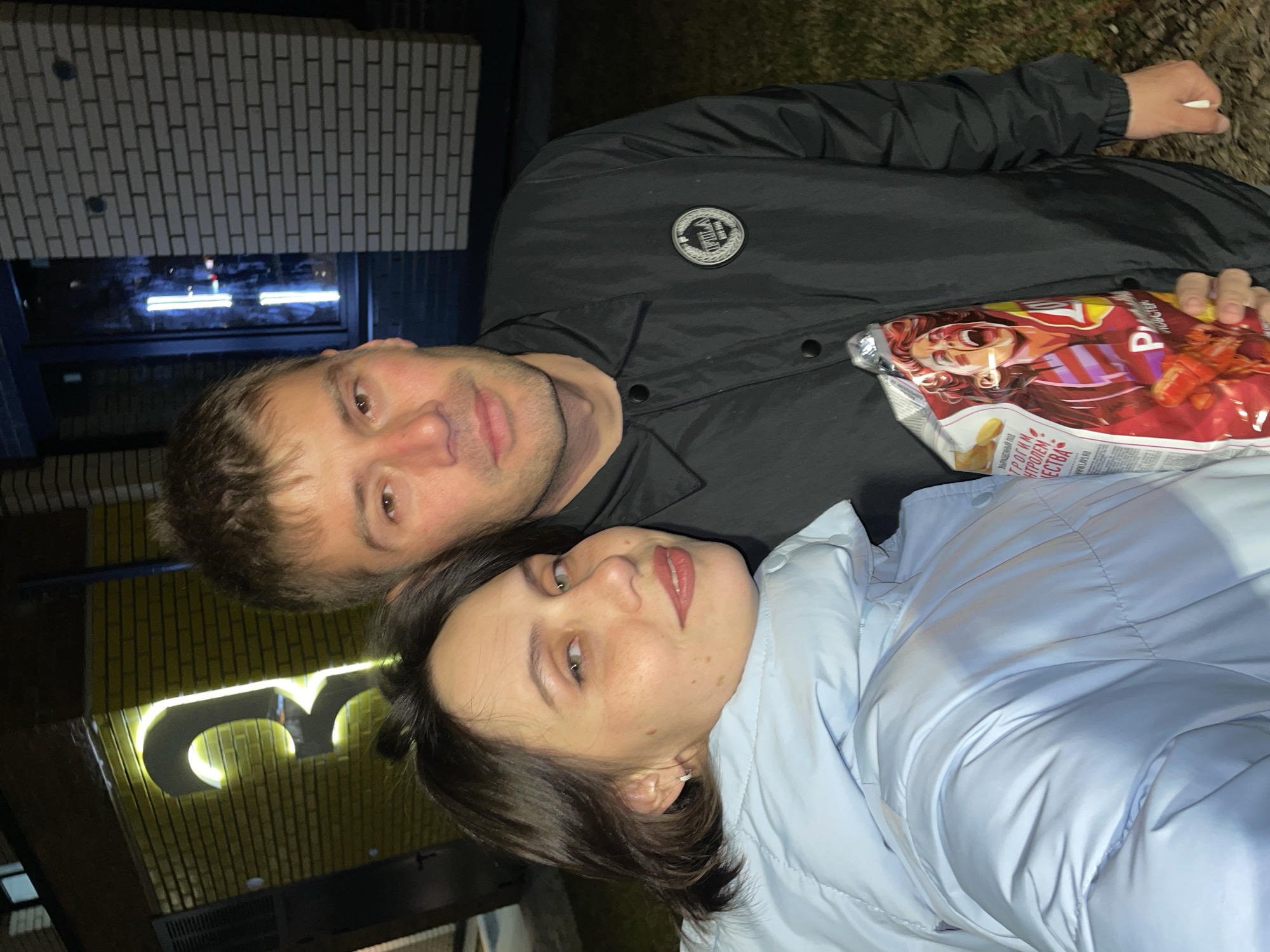 A man and a woman are posing for a photo outdoors, likely at night. The man is wearing a black jacket with a circular emblem on the chest. He holds a bag of chips. The woman is wearing a light blue puffer jacket. The background includes a brick building, illuminated signage in Cyrillic, and some greenery. The location is likely in Pervomaysky, Russia.