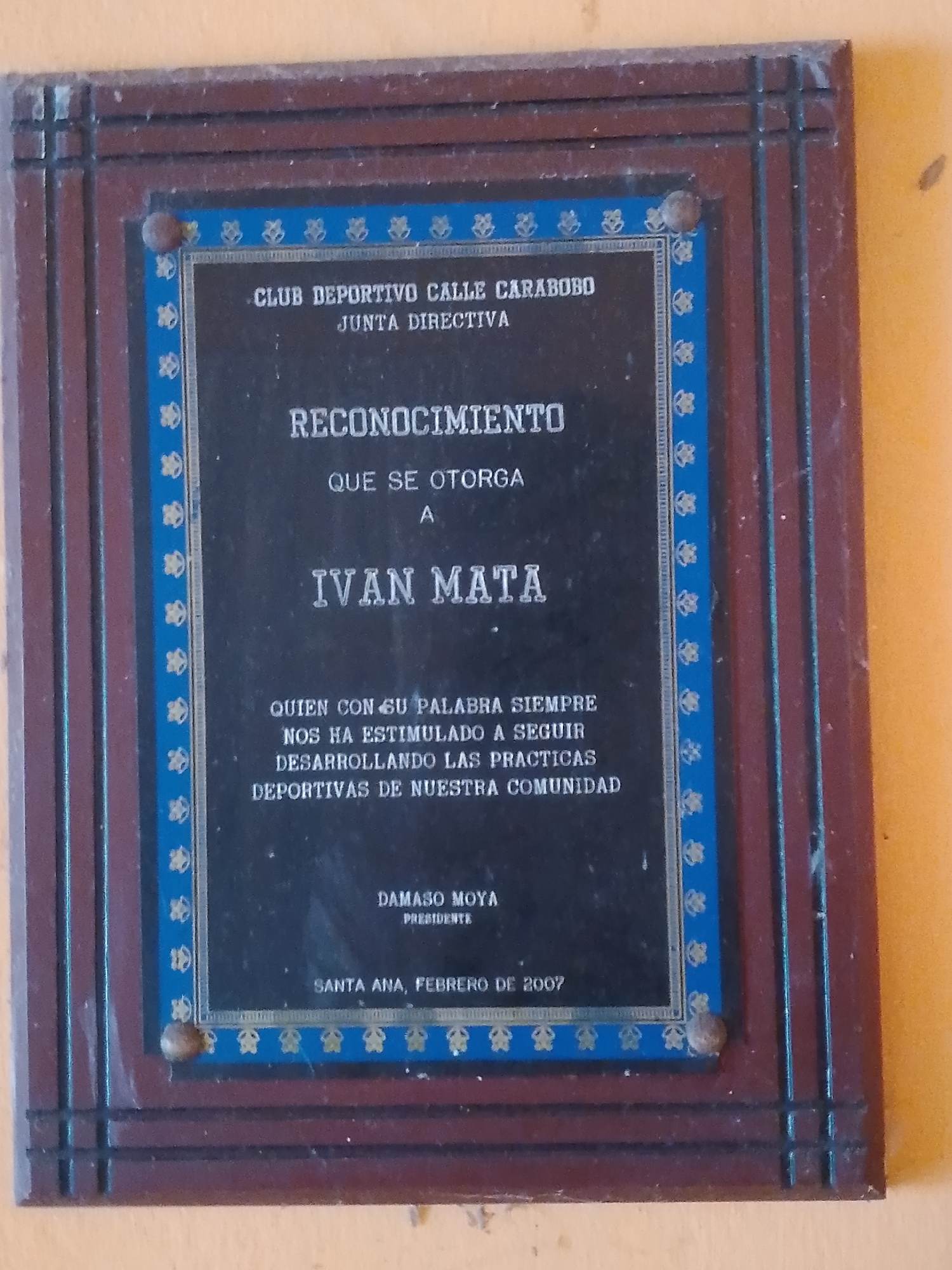 The image shows a wooden plaque with a dark blue border featuring a repeating floral pattern in light blue and yellow. The plaque is mounted on a wall. The text on the plaque is in a white, serif font. The top of the plaque reads "CLUB DEPORTIVO CALLE CARABOBO JUNTA DIRECTIVA". Below this, it says "RECONOCIMIENTO QUE SE OTORGA A IVAN MATA". Further down, it reads "QUIEN CON SU PALABRA SIEMPRE NOS HA ESTIMULADO A SEGUIR DESARROLLANDO LAS PRACTICAS DEPORTIVAS DE NUESTRA COMUNIDAD". At the bottom of the plaque, it states "DAMASO MOYA PRESIDENTE" and "SANTA ANA, FEBRERO DE 2007". The plaque is framed by a dark brown wooden border with grooved detailing. The wall behind the plaque is painted in a light yellowish-orange color. The overall lighting suggests an indoor environment.
