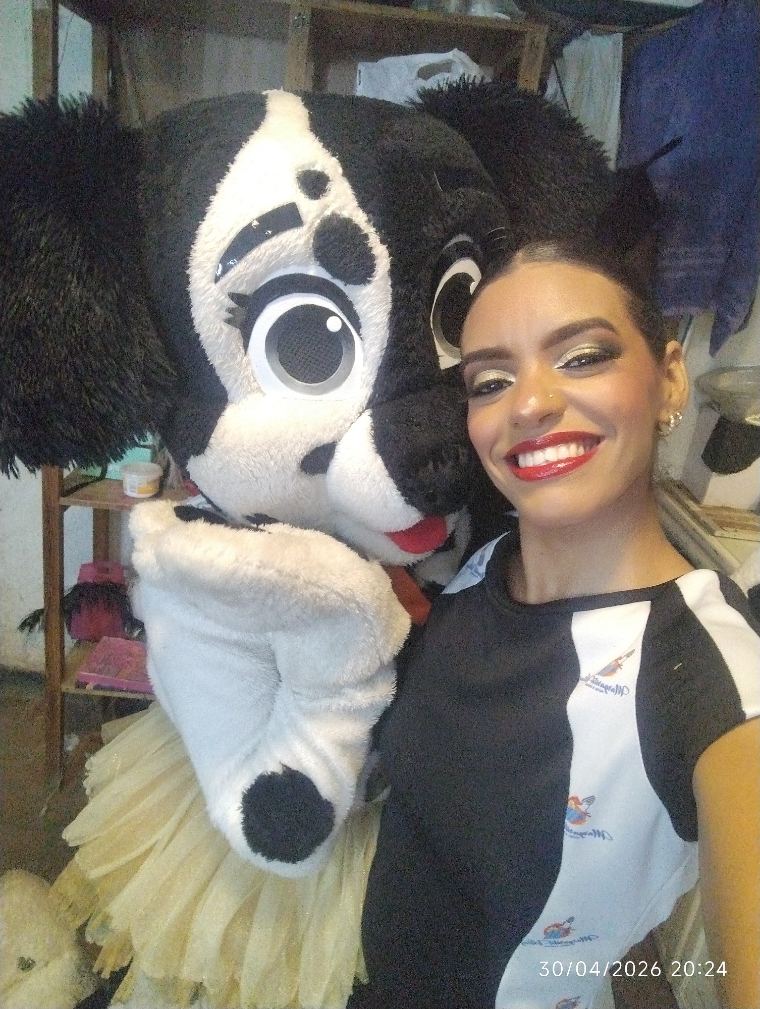 A woman with dark hair pulled back and striking makeup, featuring gold eyeshadow, dark eyeliner, and bright red lipstick, smiles broadly for a selfie alongside a large, black and white mascot costume. The woman also has a small nose piercing and is wearing a black and white short-sleeved top with a logo on the white section. The logo depicts a winged figure holding what appears to be a torch or banner, with text that includes "Servicios MP" or similar wording.
The mascot is a fluffy, anthropomorphic character, likely a dog or a similar animal, with a large head, big cartoonish eyes with prominent pupils, and a visible red tongue. Its fur is primarily black with significant white patches, particularly around its face and belly. The mascot's body appears to be wearing a yellow or gold layered, ruffled skirt-like garment. The scene takes place indoors, possibly in a backroom or storage area in Porlamar, Venezuela, given the cluttered background featuring wooden shelves holding various items like jars and fabric. The overall impression is one of a cheerful interaction or promotional event. A digital timestamp in the bottom right corner of the original image reads "30/04/2026 20:24".
