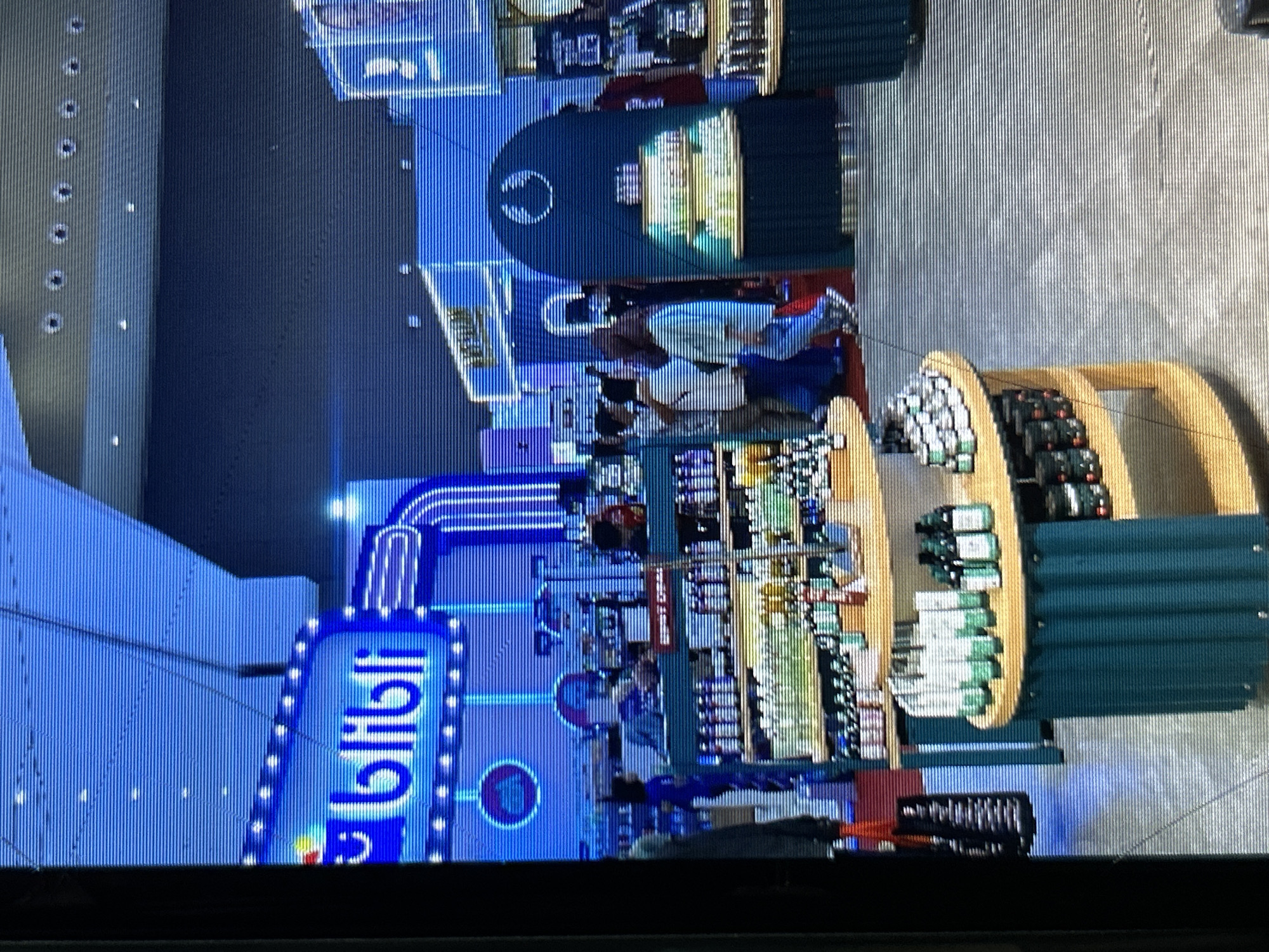 The image shows an interior view of a brightly lit retail space, likely a department store or exhibition booth, with a prominent blue neon sign that reads "bilibili". The sign features a cartoonish character to the left of the text and is surrounded by illuminated bulbs. In the foreground, there are several display shelves stocked with various products, including bottles that appear to be cosmetics or personal care items. Some shelves are arranged in a circular fashion. People are visible browsing the merchandise and interacting within the store. The environment is characterized by a modern aesthetic with a predominantly blue and teal color scheme, suggesting a contemporary retail or promotional setting. The flooring is a neutral-toned tile or polished concrete. This scene is likely located in a shopping mall or exhibition center in Depok, Indonesia, given the context of the query.
