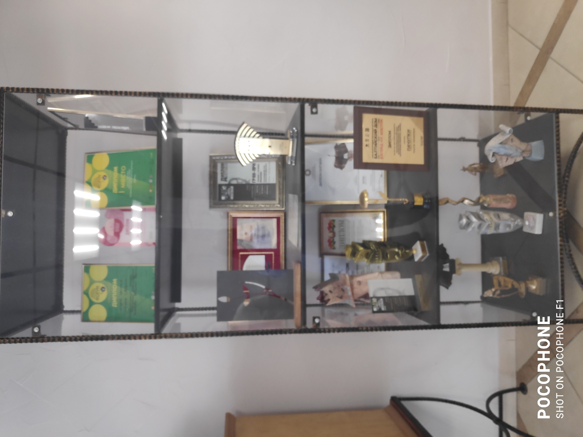 The image shows a glass display case filled with various awards, certificates, and trophies. The location is unknown as there are no identifiable city features, people, or environmental clues. The display case is multi-tiered and houses a collection of items signifying achievement or recognition.