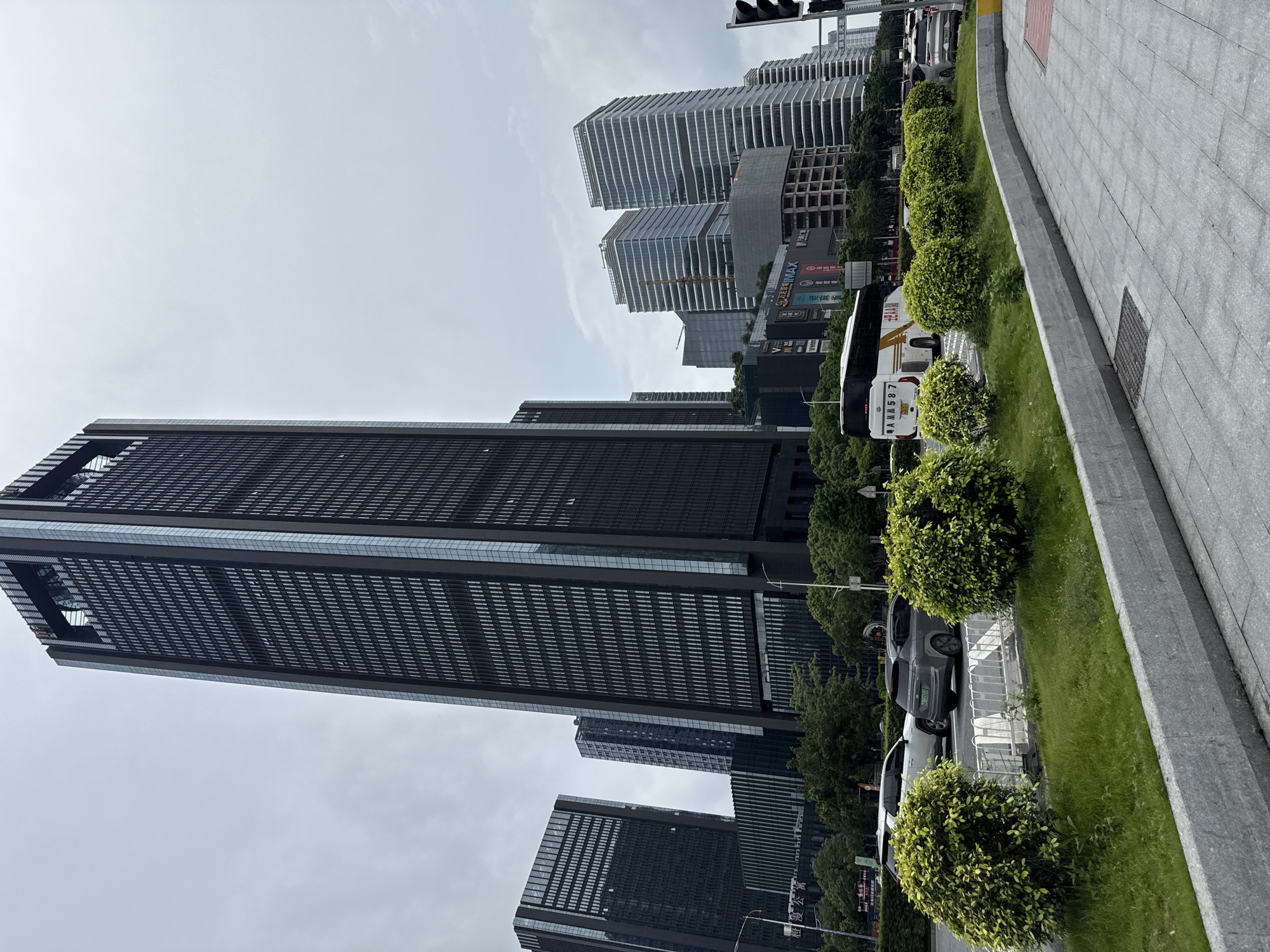 The image shows a street view in Haizhu District, China, with several modern skyscrapers dominating the skyline. The buildings are tall and made of glass and dark materials, reflecting the overcast sky. In the foreground, there is a road with cars and a bus. Lush green trees and manicured bushes line the road. The sidewalk is paved with gray tiles. Traffic lights are visible on the right side of the image. The scene appears to be a typical urban environment, possibly during the daytime, given the lighting conditions. There are no people clearly visible in the image.