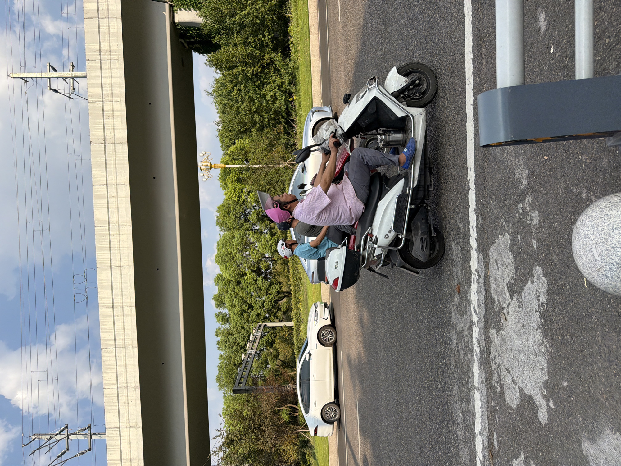 In Jiangjin, China, on a sunny day, a scooter carries two people past a white sedan. The scooter, a white electric model with a storage box on the back, is being driven by a man wearing a pink checkered shirt, grey pants, blue shoes, and a pink helmet. Behind him, a person wearing a blue shirt and a white helmet sits on the scooter. The white sedan is parked on the side of the road. The scene is set against a backdrop of green trees and a cloudy sky. An elevated structure, possibly a highway overpass, runs across the top of the image, with power lines extending from poles.