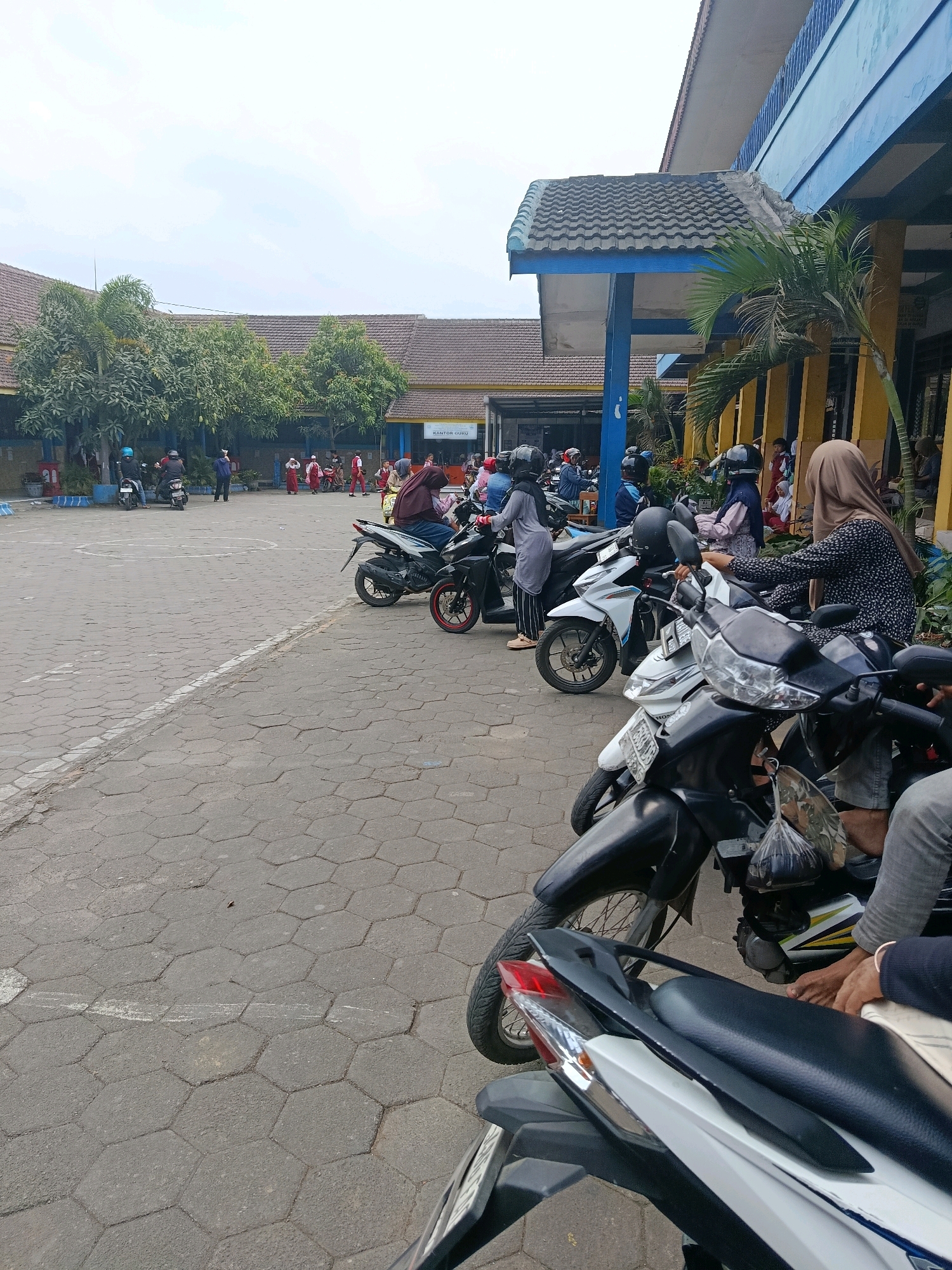 The image depicts an outdoor scene in front of a school building in Bululawang, Indonesia. The foreground is dominated by several parked motorcycles, with parts of at least three visible: a white and black motorcycle in the immediate foreground, a white motorcycle to its left, and a black motorcycle further to the left. Several people are seated on or standing near the motorcycles. One person in a brown hijab is seated on the white motorcycle, facing away from the camera. To the left of this person, another person in a grey hijab is standing next to a motorcycle.
In the middle ground, more motorcycles are parked, and several individuals, some wearing school uniforms, are seen walking and standing. A few trees and a palm tree are visible to the left and right of the building. The building itself has a blue facade with yellow pillars and a brown tiled roof. A sign above an entrance reads "KANTOR GURU" (Teacher's Office). The ground is paved with hexagonal interlocking bricks. The background shows more trees and the sky with scattered clouds. The overall impression is a schoolyard with students and staff present, possibly during a break or at the end of the school day, with a concentration of motorcycles suggesting a common mode of transport.