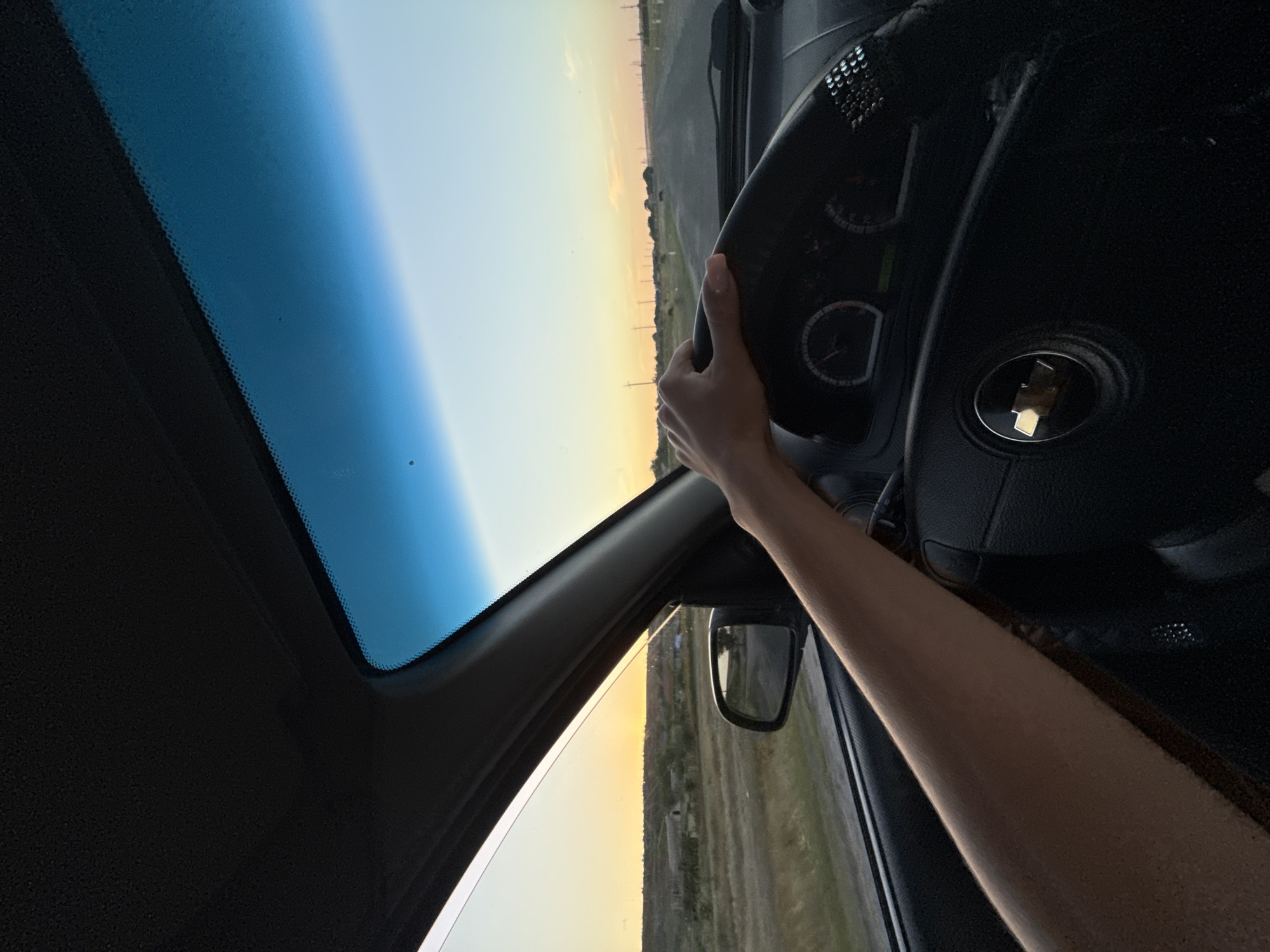 The image shows a first-person perspective of someone driving a car on a road during sunset. The driver's arm and hand are visible, resting on the steering wheel. The steering wheel has a Chevrolet logo in the center. The dashboard with gauges is also visible. Outside the car, a rural landscape with fields and some buildings can be seen. The sky is a gradient of blue and orange, indicating the sun is setting. The road ahead appears to be paved and stretches into the distance. The location is specified as Cioc-Maidan, Moldova.
