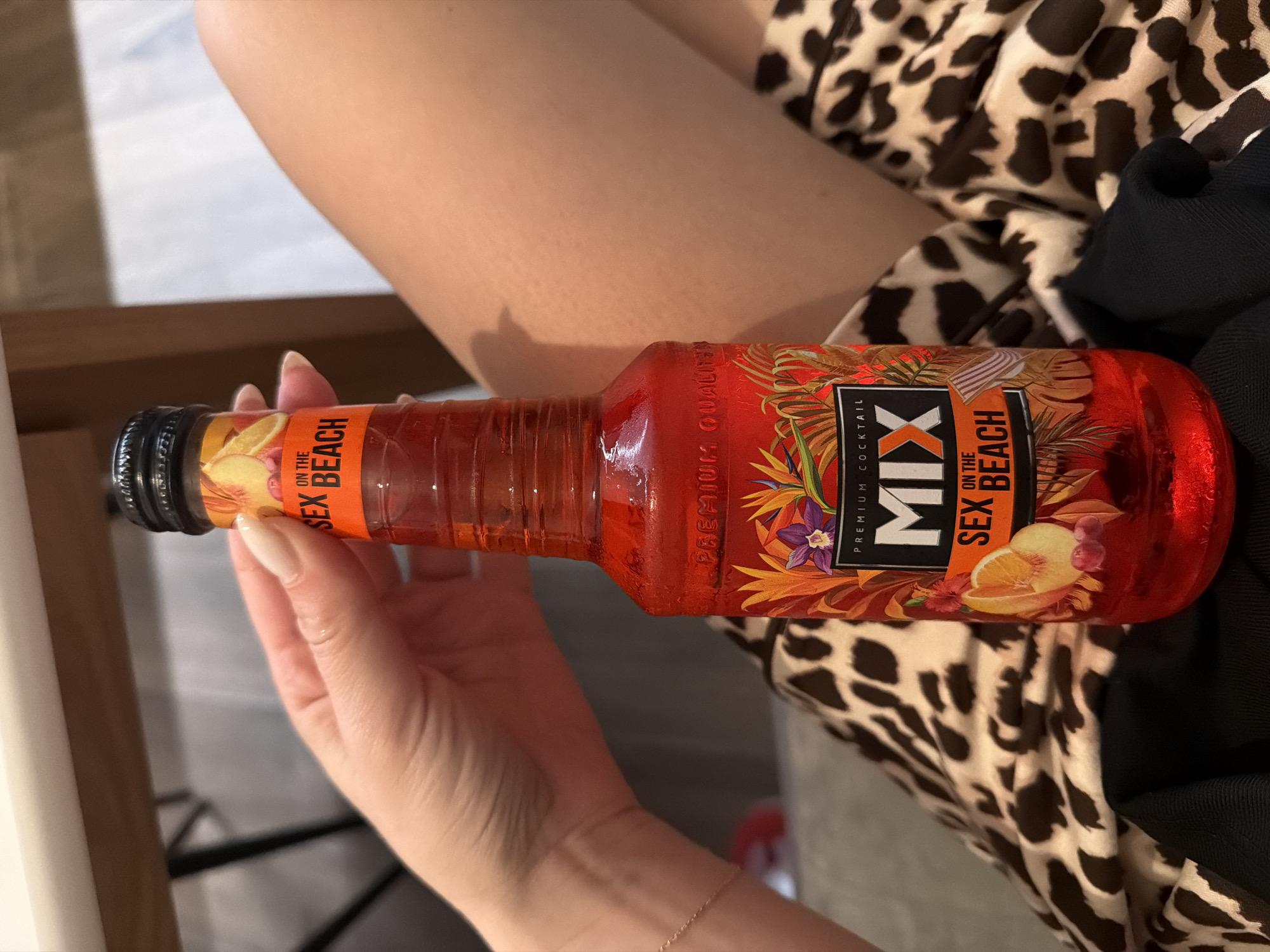 A person is holding a bottle of "Mix Sex on the Beach Premium Cocktail". The person is seated, as their upper arm and upper part of their leg are visible. They are wearing a brown and white animal print garment and a black garment is also partially visible. The location is unknown.