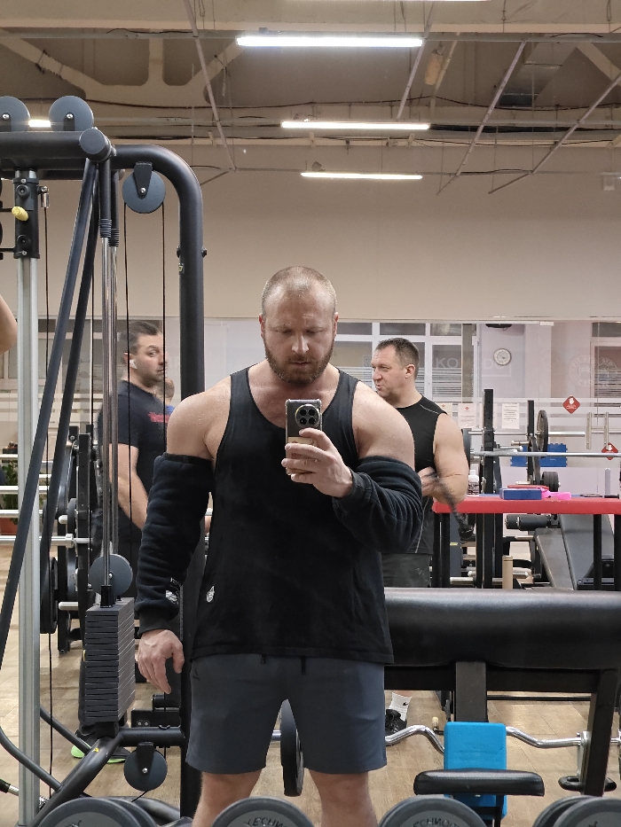 A male individual with a bald head and a reddish-brown beard is depicted in a mirror selfie within a gymnasium. He wears a black tank top, dark grey shorts, and black full-length compression arm sleeves. He holds a smartphone with a gold-toned case in his right hand, positioning it to capture his reflection.
In the reflection behind the primary subject, two other males are visible. To the left, partially obscured by gym equipment, a male with dark hair wearing a black t-shirt is visible. To the right, another male with short dark hair, wearing a black sleeveless shirt, stands with his arms crossed.
The environment is a brightly lit gym, indicated by overhead fluorescent strip lights and light-colored flooring. Various gym equipment is present, including a cable machine with weight stacks on the left, dumbbells (some with "TECHNOGYM" branding) on the floor in the foreground, and a weight bench with a red pad and a barbell rack in the background to the right. A white-faced clock is visible on the back wall. Text on a white wall, visible in the mirror's reflection, includes "СПОРТКЛУБ" (Sports Club) and "ЧАЙКОВСКИЙ", confirming the location as a sports club in Chaikovsky, Russia.