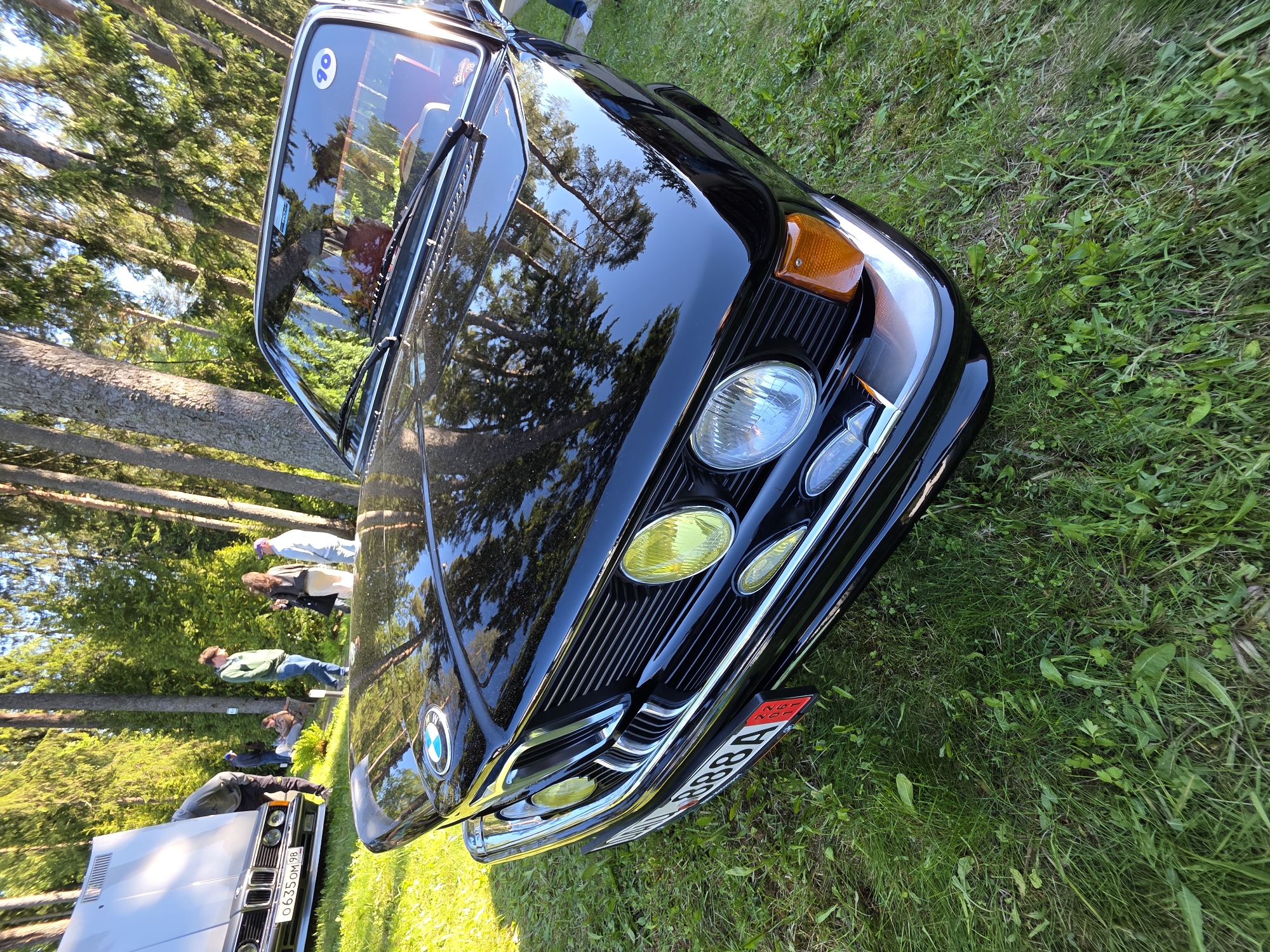The image shows a close-up view of a black BMW automobile parked on a grassy field in Repino, Russia. The front of the car is prominently featured, including its headlights, grille, and bumper. The car’s hood reflects the surrounding trees. A group of people stand nearby in the background, observing the cars. Another car is partially visible in the lower left of the frame, displaying its front grille and license plate. The environment suggests a park or similar outdoor setting.