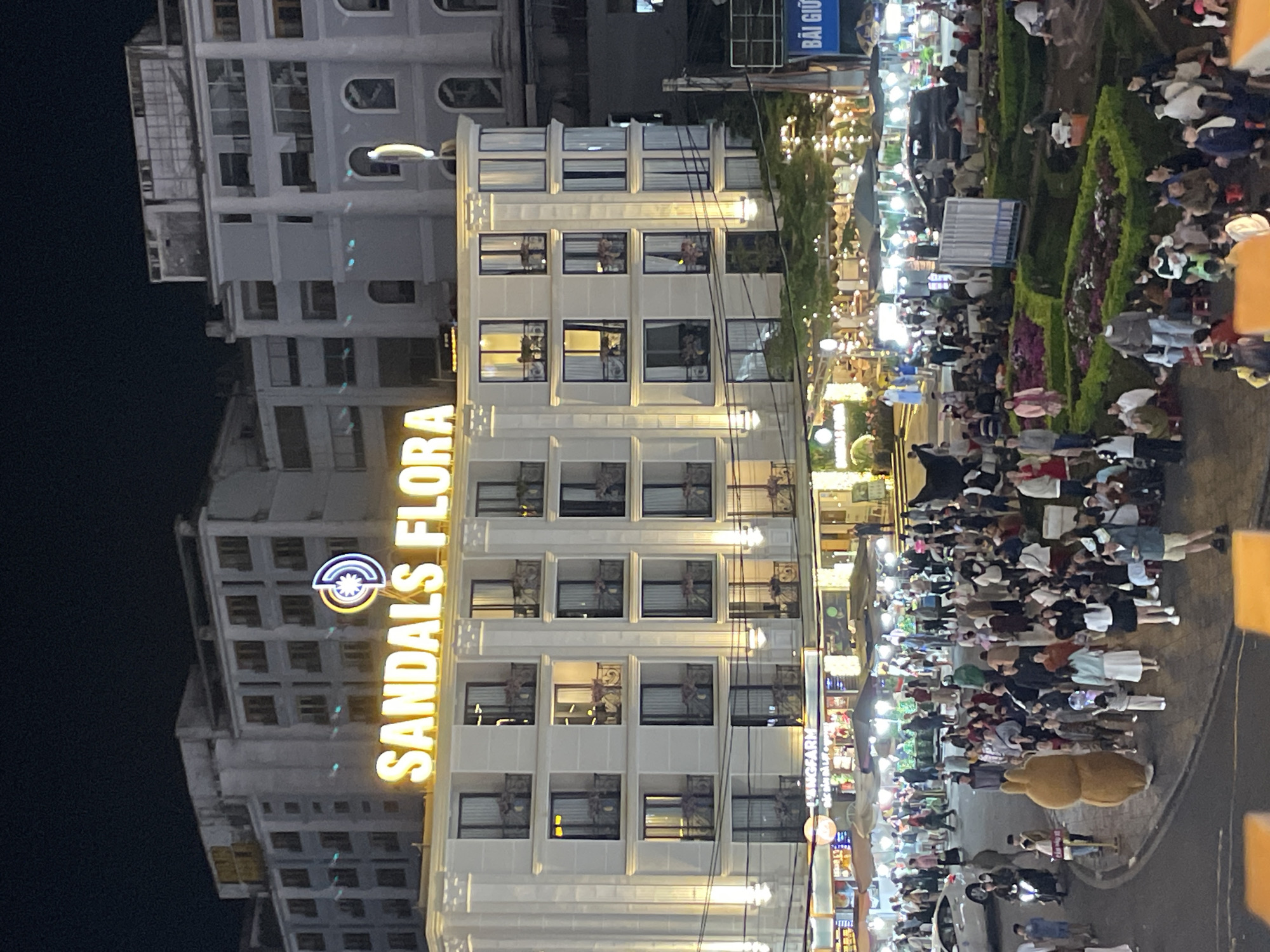 The image depicts a bustling street scene at night in Da Lat, Vietnam, likely near Xuan Huong Lake, a popular tourist destination. A brightly lit building with the neon sign "SANDALS FLORA" is prominent. The building has multiple floors with balconies and illuminated windows, suggesting it is a hotel or commercial establishment.
Below the building, a lively night market is in full swing. Numerous stalls are set up, illuminated by strings of lights, creating a festive atmosphere. A large crowd of people, appearing to be tourists and locals, are milling about, browsing the market, and enjoying the evening. Some individuals are sitting on the ground, while others are standing and interacting.
In the foreground, a person in a large, brown, cartoon-like costume, possibly a mascot, stands out among the crowd. A white car is parked on the left side of the image, and a motorcycle can be seen in the middle-left. A blue sign with Vietnamese text is visible on the right side of the image. The ground appears to be paved, and there are landscaped garden areas with colorful flowers and green bushes. The overall impression is one of a vibrant and active urban night scene in Da Lat.