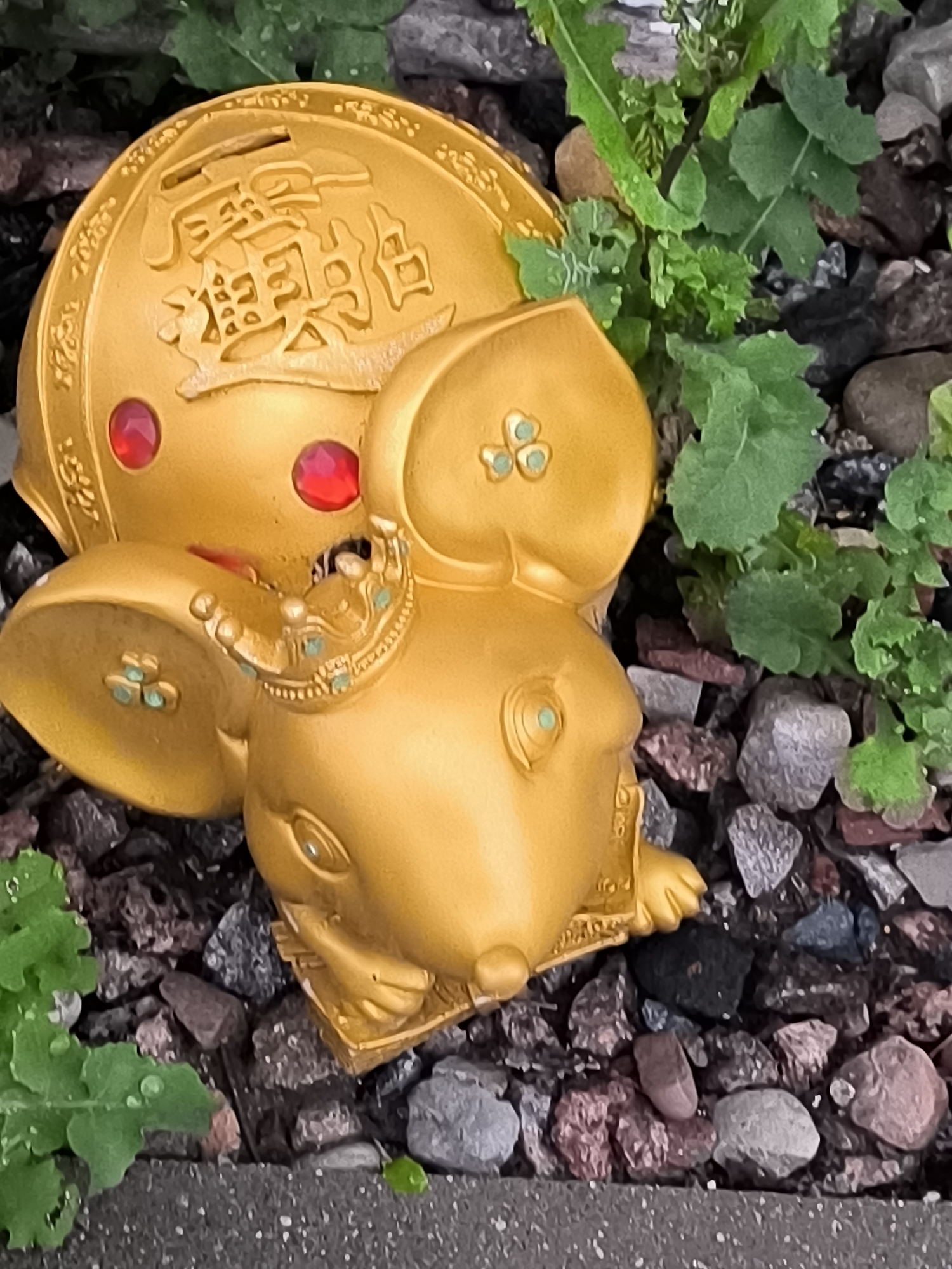 The image shows a close-up, high-angle shot of a golden mouse statue placed outdoors on a bed of dark and light-colored gravel and stones. Green leafy plants are growing among the rocks and partially obscure the statue. The statue is ornate, featuring red circular embellishments on its head, and a small crown adorned with green jewels on its ear. Chinese characters are embossed on the top of the statue's head. The lighting suggests it is daytime. The image composition focuses on the statue and its immediate surroundings.