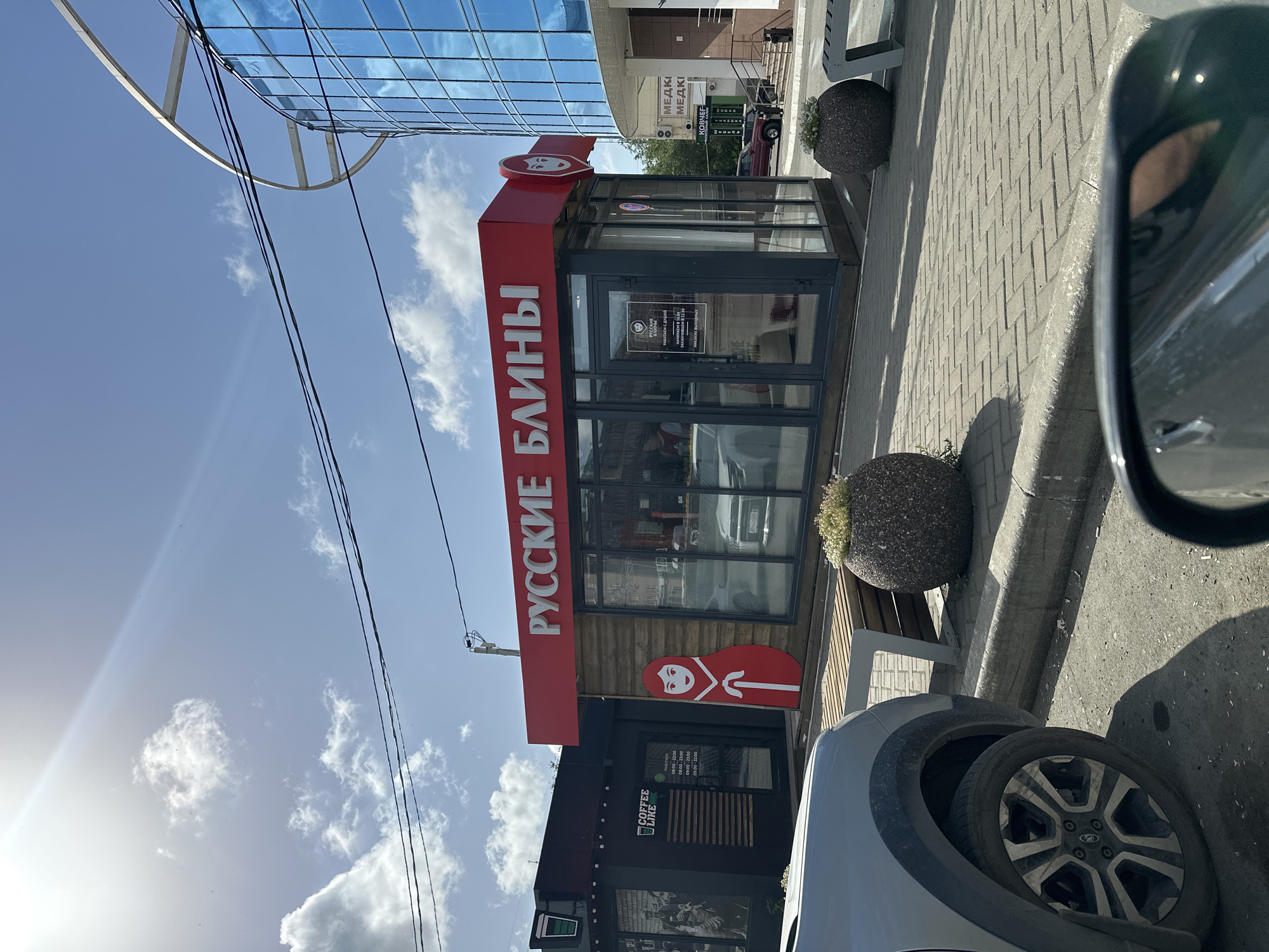 The image depicts a sunny daytime scene in Orsk, Russia, likely on a street or plaza. In the foreground, the left side of a silver SUV is visible, with its wheel and part of the fender in focus. To the right, a portion of a black car's side mirror reflects the surroundings.
The main focus of the image is a small retail kiosk with a bright red sign above it that reads "РУССКИЕ БЛИНЫ" (Russian Blini). The kiosk has large glass windows and a wooden facade. A large, stylized cutout of a traditional Russian nesting doll (Matryoshka) stands to the left of the kiosk entrance. Inside the kiosk, a person is partially visible behind the counter. Two round, stone planters with greenery sit on the paved walkway in front of the kiosk. Behind and to the right of the "Russian Blini" kiosk, a modern building with a blue glass facade and a curved metal structure is partially visible. Further to the right, a medical clinic sign "МЕДКИ" is present. Adjacent to the "Russian Blini" kiosk on the left is another smaller black kiosk with a "COFFEE LIKE" logo and its operating hours displayed on the window. The ground is paved with interlocking bricks, and a concrete curb separates the walkway from the street. The sky is blue with scattered white clouds and visible power lines.