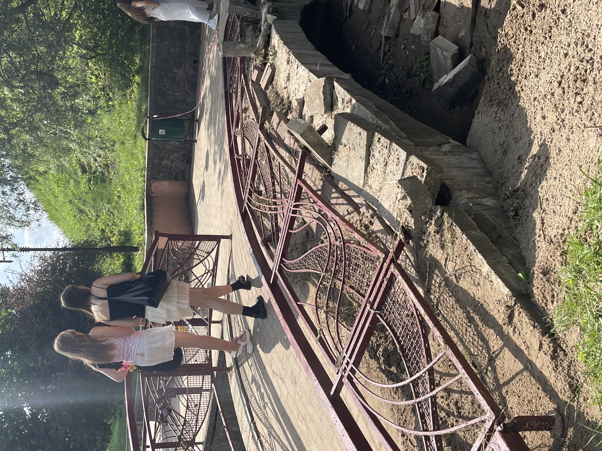 The image depicts a damaged bridge railing, likely in Hrodna, Belarus. Two women are walking away from the camera on the intact portion of the bridge, while the foreground shows the collapsed railing and surrounding debris. The bridge appears to be made of concrete and has decorative metal railings. The area around the bridge is sandy, with some grass and trees visible in the background. A green trash can and caution tape are also present. The damage to the bridge suggests recent structural failure.