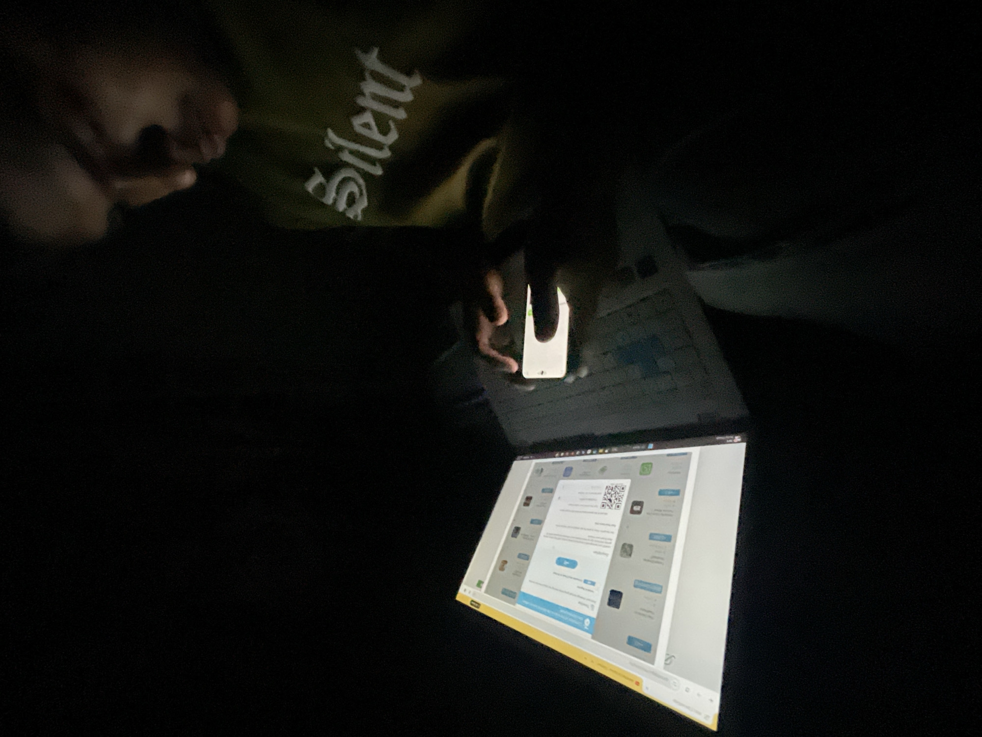 A person is holding a smartphone and scanning a QR code on a laptop screen in a dark environment. The laptop screen displays a webpage with a QR code. The person is wearing a garment with the word "Silent" on it. Location information (Dhaka, Bangladesh) is not visible in the image.
