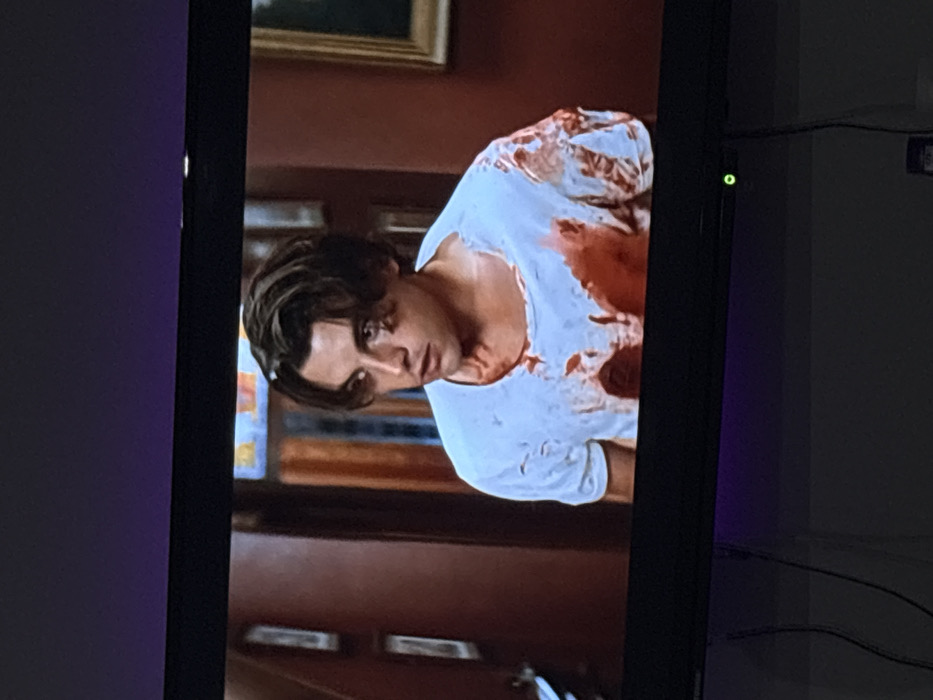 The image displays a photograph of a television screen showing an indoor scene. The specific location in the city of Ботево, Bulgaria, cannot be determined from the visual content presented.
On the television screen, a young adult male is depicted from the chest up, positioned slightly right of center. He has medium-length, wavy dark brown hair, fair skin, and dark eyes, looking intently to his left (viewer's right) with a serious or concerned expression. He is wearing a white t-shirt, heavily saturated with large, dark red stains consistent with blood, primarily across the chest, left shoulder, and stomach area, with smaller splatters on the right arm. The fabric of the t-shirt appears distressed, particularly around the neckline.
The background behind the male subject features an interior setting with dark reddish-brown walls and dark wooden architectural elements, possibly door frames or panels. On the left wall, two vertically oriented, rectangular framed objects are visible. On the right, a section of a framed painting with a gold-colored frame is partially discernible. Further back, a decorative glass or stained-glass window with geometric patterns in shades of yellow and blue is visible.
Below the television screen, a black flat-screen TV bezel is visible, featuring a small green indicator light on its lower right side, indicating power. Below the bezel, dark cables descend against a plain, light-colored wall. A subtle purple ambient light glow is present on the wall below the television. No direct interactions or specific actions other than the male subject's focused gaze are observable within the depicted scene on the screen. The presence of significant bloodstains implies prior violent activity or injury.