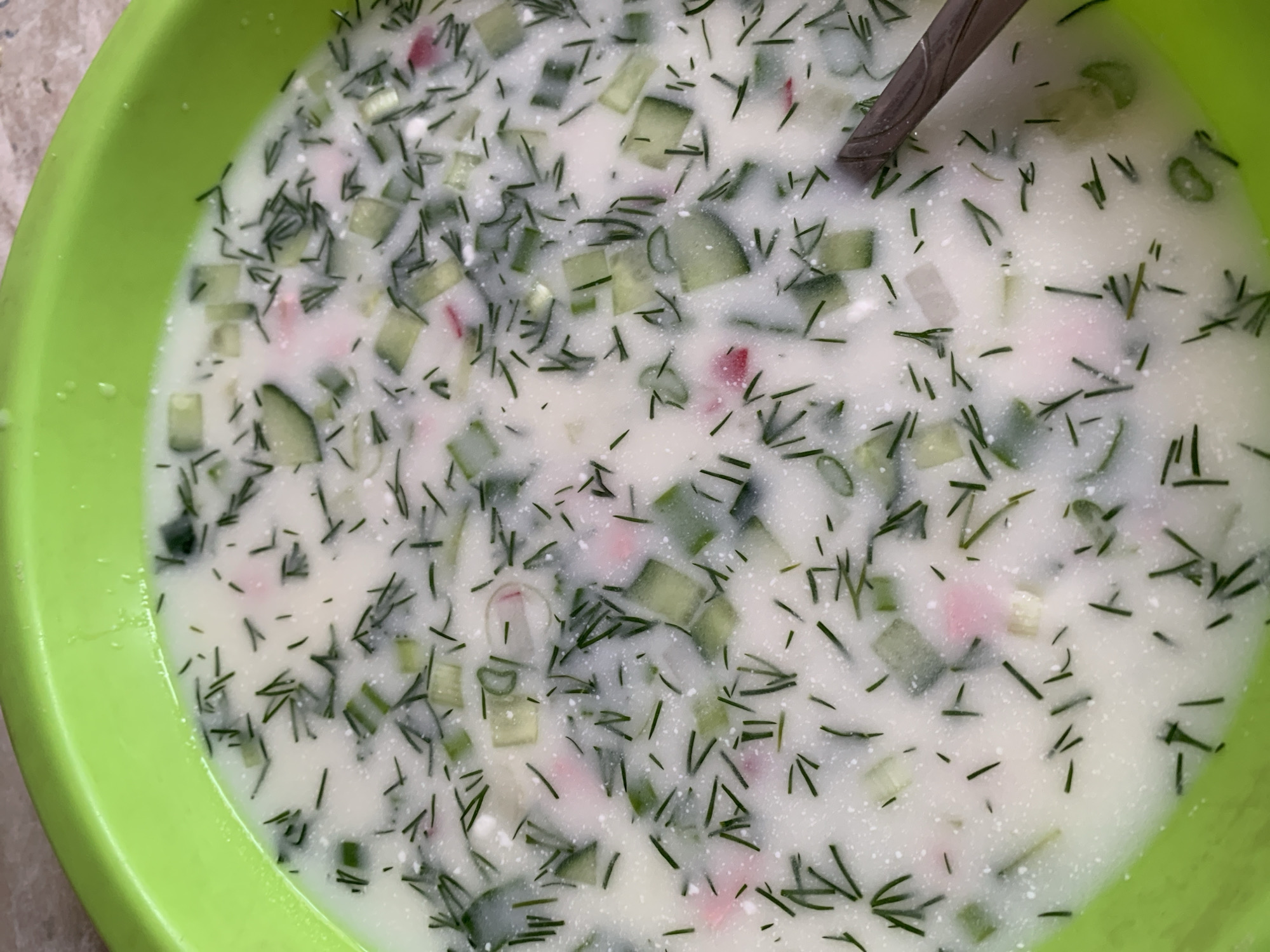 A close-up overhead shot shows a large, green bowl filled with a cold soup called okroshka. The soup has a white, milky base with chopped cucumbers, dill, radish and perhaps other ingredients visible throughout. A gray spoon rests inside the bowl. The location appears to be indoors. The city is unknown.