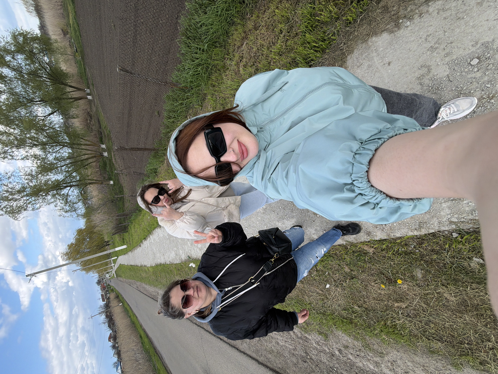 The image shows three women taking a selfie outdoors on what appears to be a path or road. The woman in the foreground is wearing a light blue hooded jacket and dark sunglasses. Behind her and to the left, another woman is wearing a beige jacket and sunglasses, making a peace sign with her hand. The third woman, closer to the bottom of the frame, is wearing a black jacket and sunglasses and also making a peace sign. The background features a road, some green vegetation, fields, and a partly cloudy sky. The location appears to be rural, likely a roadside area.
