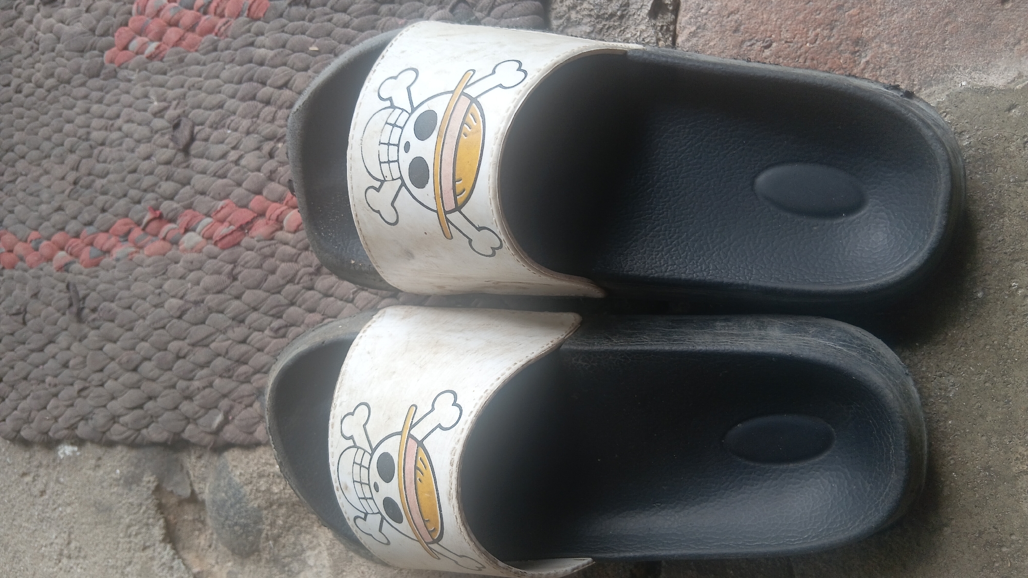 Two black slide sandals with white straps featuring a Jolly Roger-style skull with a hat and crossed bones are placed side-by-side on a textured gray and pink mat. The sandals appear worn, with some dirt and scuff marks on the straps and soles. The background consists of a rough, textured surface resembling concrete or pavement. The image is taken from a top-down perspective. The location is identified as Prabumulih, Indonesia.