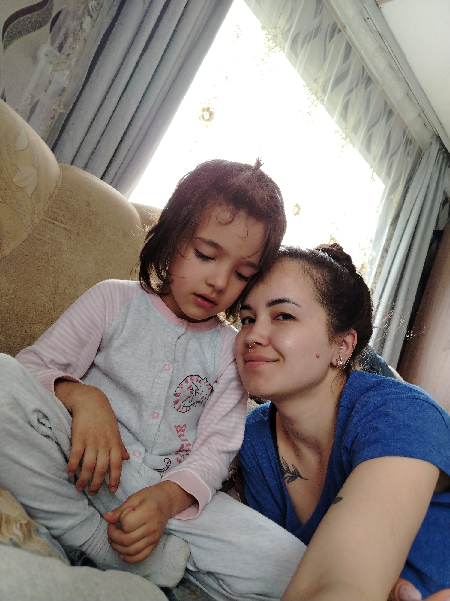 A woman with dark hair pulled into a bun is sitting closely with a young child in pajamas. The woman is wearing a blue t-shirt and has multiple piercings in her ears and a nose ring. A tattoo is visible on her collarbone. The child is wearing pink and grey pajamas with a cat design. They are sitting on a beige sofa in front of a window with lace-trimmed curtains. The image was taken in an indoor environment, potentially a home. The location is Гожан, Russia.
