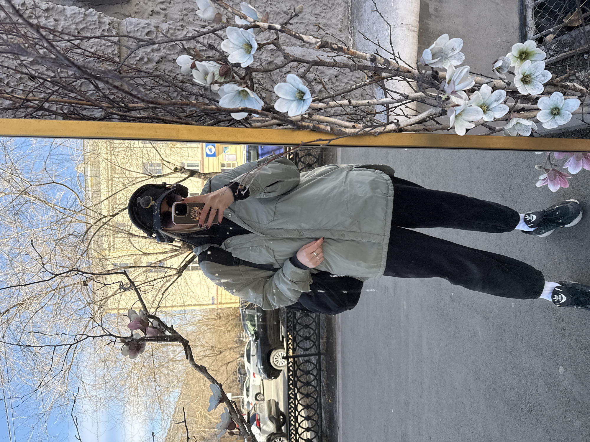 A woman is photographed in a mirror with a gold frame. The mirror is leaning against a building in Chita, Russia. On top of the mirror, there are branches with white flowers. In the reflection, the woman is wearing a helmet, sunglasses, a light green jacket, and black pants with black and white shoes. She is holding a phone and taking the picture. The mirror also reflects a street, a building, a fence, and cars. The sky is visible through the branches of a tree.