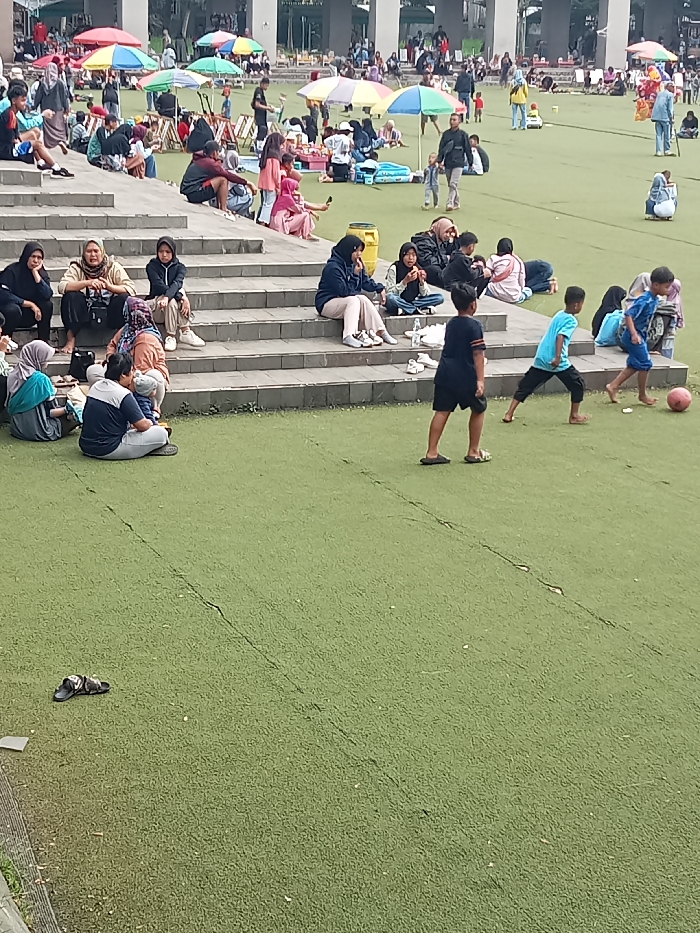 This media file depicts a lively outdoor scene at a park or recreational area in Singaparna, Indonesia. The setting features a large expanse of artificial green turf and concrete steps that serve as tiered seating.
Numerous people are present, ranging from children to adults, suggesting a family-friendly gathering or a public holiday. On the concrete steps, various groups are seated, many women wearing hijabs, observing the activities or socializing. In the foreground, a man in a blue shirt and gray pants is seated on the lowest step next to a woman in a light blue hijab.
The central activity involves three young boys playing soccer on the artificial turf with a red ball. One boy, barefoot and in a light blue shirt, is positioned near the ball, while another boy in a black shirt and flip-flops stands a short distance away. A pair of black sandals are visible lying on the turf in the lower-left corner, likely discarded by one of the players or a spectator.
In the mid-ground and background, more people are scattered across the green space. Many are gathered under an array of colorful umbrellas (red, blue, green, yellow, and multi-colored), indicating a sunny day and a need for shade. Some individuals are walking across the field, and others appear to be having picnics or relaxing. The background features some indistinct building structures with tall pillars, suggesting a public facility.
The overall atmosphere is casual and relaxed, indicative of leisure and community engagement. The scene is brightly lit, confirming it is daytime. No discernible text is visible in the image.