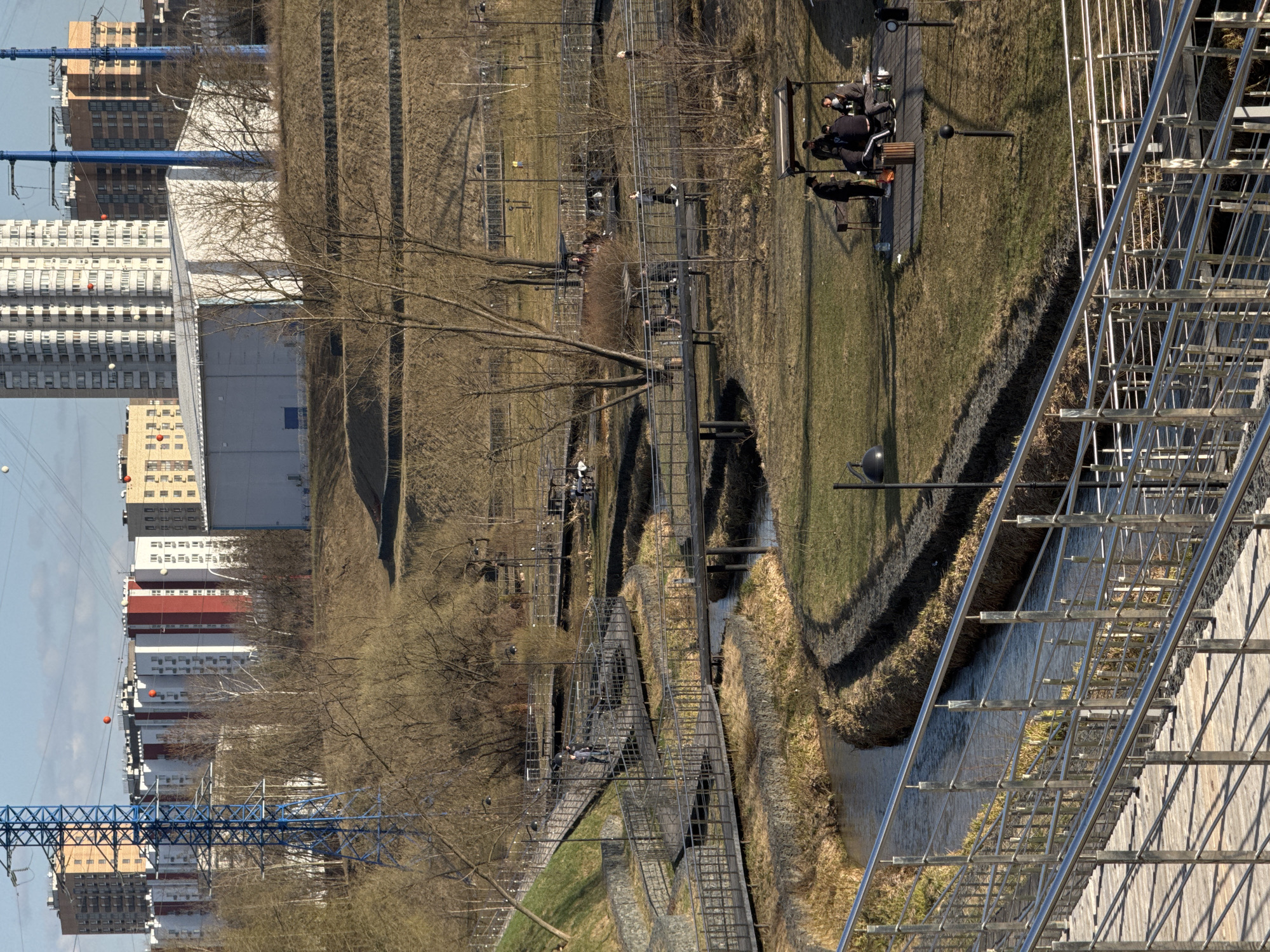 The image depicts Zaryadye Park in Moscow, Russia, showcasing an urban landscape with a mix of natural and man-made elements. In the foreground, a metal walkway with railings extends into the frame, providing a vantage point over a stream or canal. A grassy area is visible, along with pathways winding through trees. In the background, high-rise buildings and residential structures are visible, indicating an urban setting. Several people are visible throughout the park, some sitting on benches. The scene is bathed in natural daylight with visible shadows, suggesting a clear day.