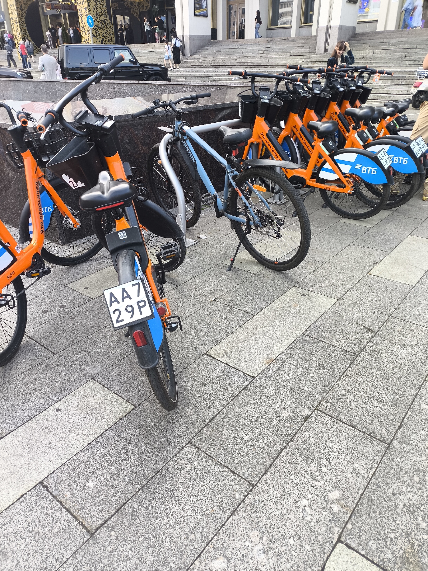 In an outdoor urban setting, a row of parked orange and blue bicycles with black seats and handlebars stands on a paved area of grey and beige granite slabs. The bicycles are oriented towards the right of the frame, with their front wheels partially visible. Each bicycle has a black basket mounted on the handlebars and a rear fender. Some bicycles display license plates, such as "AA7 29P" on one orange bicycle. There are also QR codes visible on some of the bicycles. The background shows a paved area with stairs leading up to a building with a patterned facade. Several people are visible in the background, walking on the stairs and in the vicinity of the building. A black SUV is parked near the stairs.