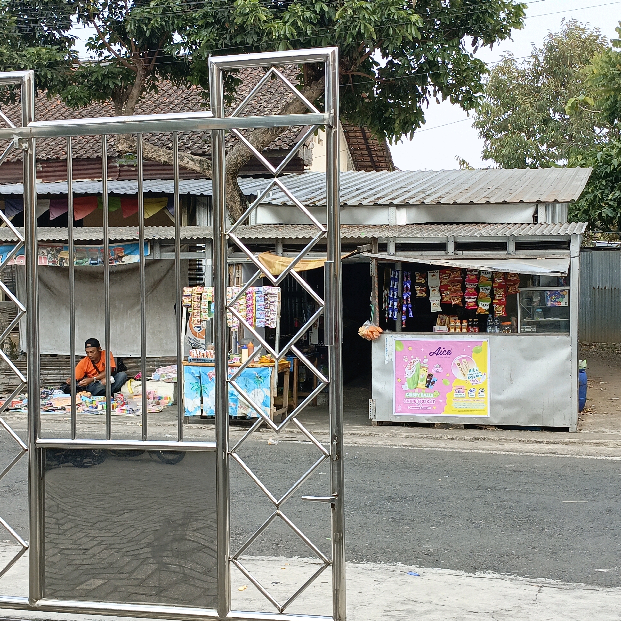 This image captures a street scene in Salatiga, Indonesia, likely in front of a small roadside shop. A man wearing an orange t-shirt and a black baseball cap is seated on the ground, surrounded by various colorful items that appear to be merchandise or toys. He is looking down and appears to be arranging or interacting with these items.
Behind the man, a small, open-air stall is visible. The stall has a corrugated metal roof and a counter displaying an assortment of packaged snacks and drinks. A large pink and yellow poster is affixed to the front of the stall, advertising "Aice Crispy Balls Shine Muscat."
In the foreground, a modern, silver metal gate with a diamond pattern is partially open, framing the scene. The gate is made of vertical bars and horizontal supports. The ground in front of the stall and to the side of the gate is paved. Trees with green foliage are visible in the background, with the distinctive red tile roofs of houses peeking out from behind. The overall impression is of a modest, everyday commercial activity on a street in Indonesia.