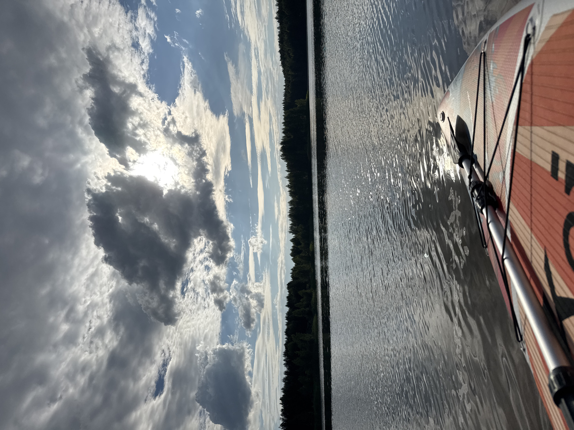 The image depicts a serene lake with a line of dark green trees along the distant shore. The water's surface is rippled and reflects the sky, which is a mix of bright blue and dramatic grey clouds, with the sun peeking through in the upper center. In the foreground, the nose of a paddleboard is visible, with a paddle resting on it. The paddleboard is orange and white with a patterned design. There are no people visible in the image. The scene suggests a peaceful outdoor setting, possibly a recreational area on a lake. The location is identified as Novyi, Russia.