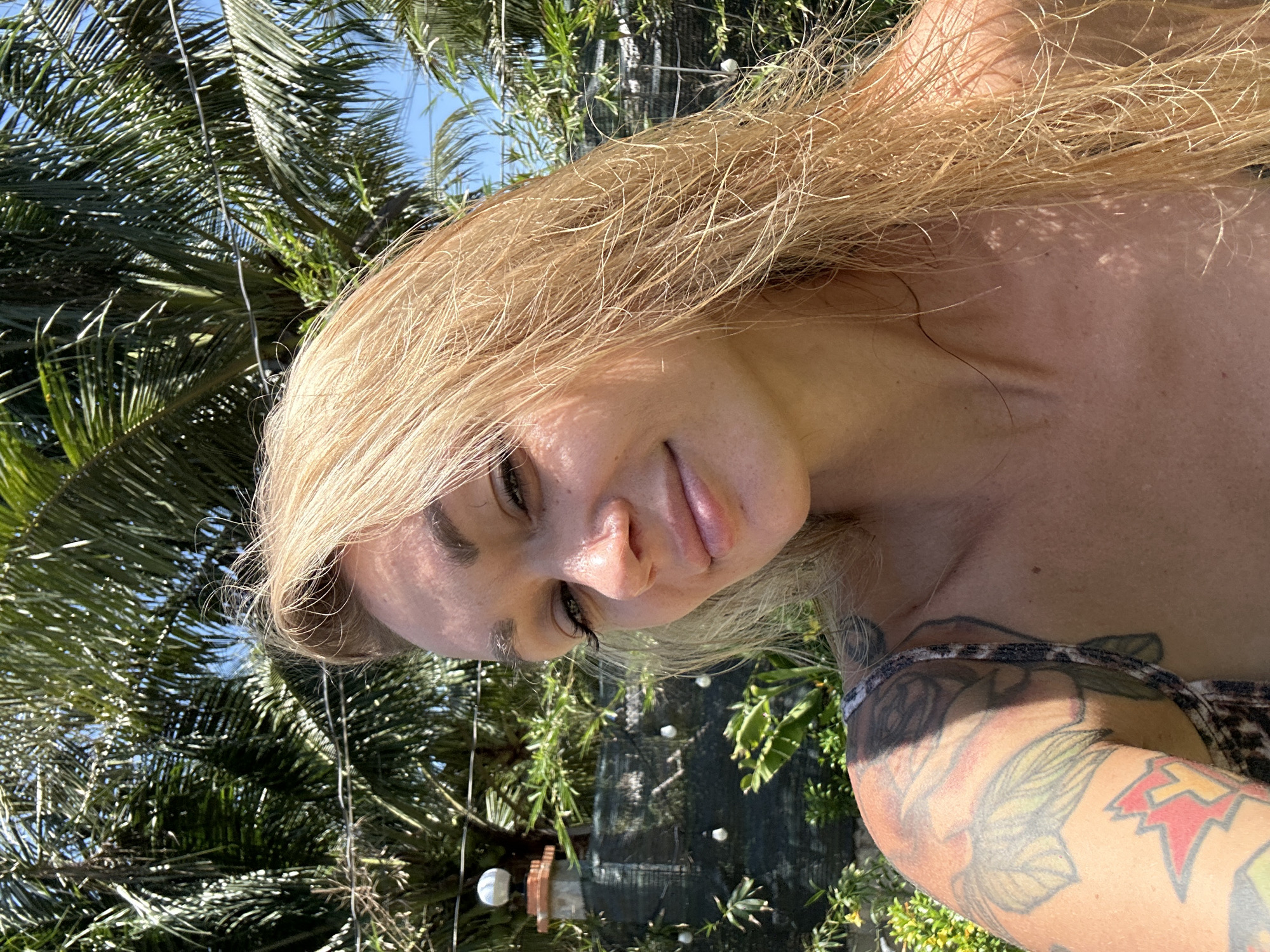 A woman with long blonde hair is pictured in a close-up shot with palm trees and other vegetation in the background, likely in Mũi Né, Vietnam. She has fair skin and a tattoo on her upper arm. A structure, possibly a fountain, is visible in the background, with string lights hanging overhead. The sky is blue and the sun appears bright.