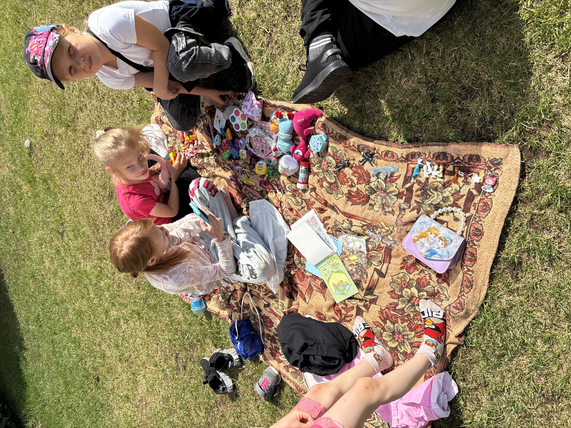 The image shows a group of children sitting on a patterned blanket in a grassy area. There are at least three girls visible, with one wearing a pink shirt and another with red hair wearing a patterned top. Another child wears a white shirt and a cap with a colorful design. Various toys and objects are scattered on the blanket, including dolls, figurines, and a small purse with a Disney Frozen image. Clothes and shoes are placed around the blanket. In the background, there is a person's leg wearing black pants and shoes. The setting appears to be outdoors on a sunny day. It is difficult to pinpoint an exact location in Izhevsk, Russia, without more contextual clues.