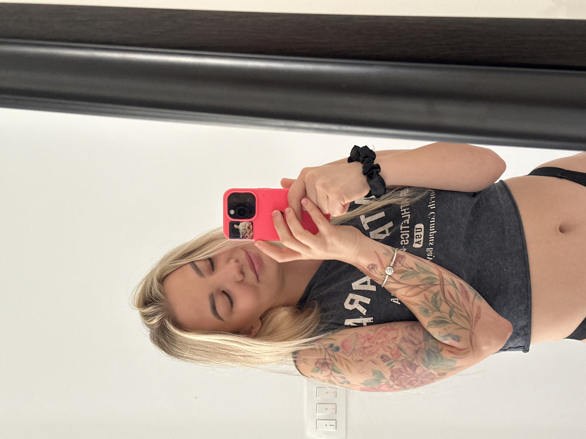 In a mirror selfie taken indoors in Budva, Montenegro, a young woman with blonde hair is captured from the mid-chest up. She has a serene expression with closed eyes and a slight smile. She is holding a bright pink smartphone with a dark case in front of her face, with the phone itself reflected in the mirror. A black scrunchie is on her right wrist, and a silver bracelet adorns her left wrist. Her left arm is extensively covered with colorful floral and foliage tattoos, visible from her forearm up to her bicep. She is wearing a dark grey cropped t-shirt, revealing her midriff, and black underwear or swimsuit bottoms. The t-shirt has reversed text on it, which appears to say "WORLD CUP". The background is predominantly white, reflecting the room's bright interior. A dark horizontal bar, likely a mirror frame or shelf, is visible at the top, and light switches are present on the lower wall. The overall setting appears clean and bright.
