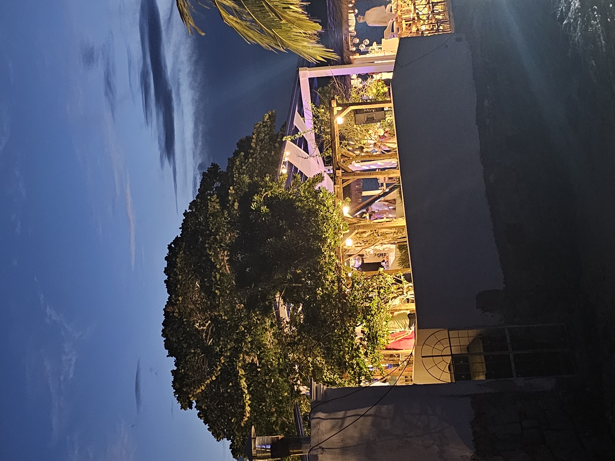 The image captures an outdoor venue in Armação dos Búzios, Brazil, taken at dusk or night. The venue appears to be a multi-level structure with wooden frames, decorated with string lights and foliage, possibly a restaurant or bar. Patrons are seated at tables, their figures indistinct. A large tree with dense green leaves partially obscures the view of the venue. The sky is visible with shades of blue and dark clouds. Part of a building with a window is visible on the right side. A palm frond is visible in the upper left corner.