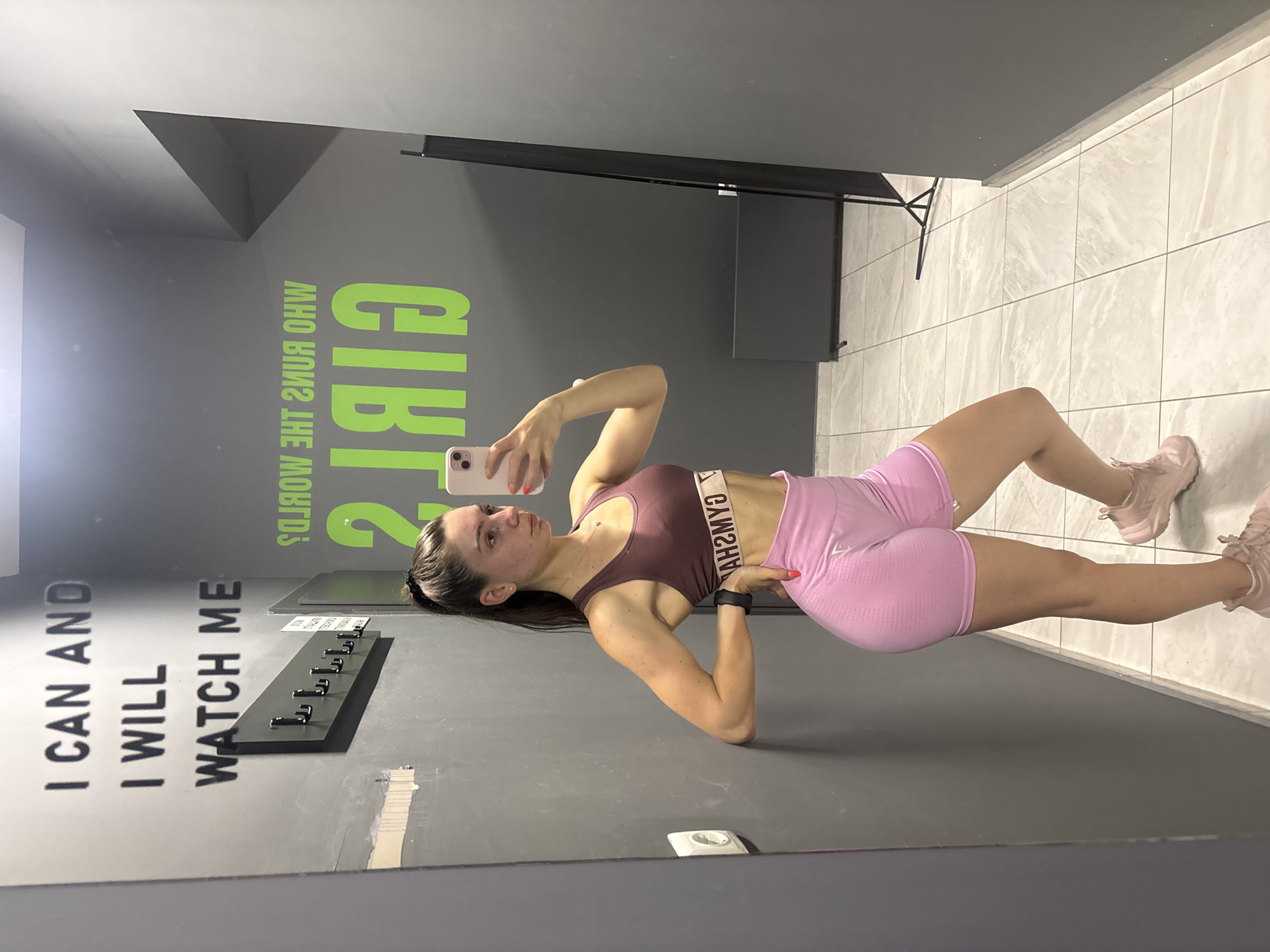 A woman in fitness attire takes a selfie in a gym setting. She wears a brown sports bra and pink shorts, with matching pink sneakers. The background includes a grey wall with motivational phrases such as "I CAN AND I WILL WATCH ME" and "GIRLS WHO RUN THE WORLD." A coat rack is visible on the wall, and the floor is tiled. The location is likely a gym in Varna, Bulgaria.
