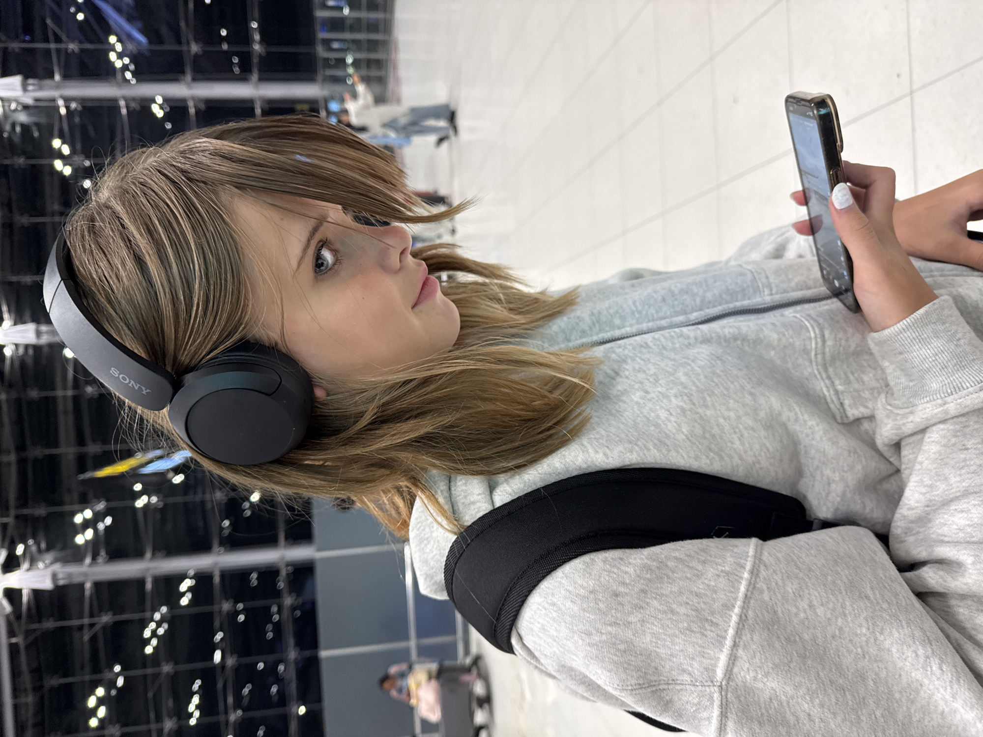 The image shows a young person wearing black Sony headphones and a grey hoodie. They are looking down at a smartphone held in their right hand. The person has blonde hair and blue eyes. In the background, there is a large, brightly lit indoor space, possibly an airport terminal, with blurred figures and luggage visible. The flooring appears to be white tile. The location is identified as Nong Prue Subdistrict, Thailand.