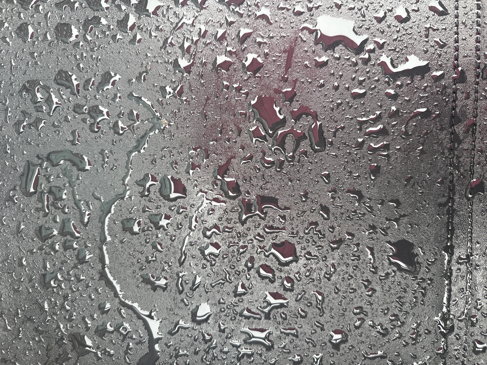 A close-up, top-down view of a textured, dark gray surface covered in numerous water droplets of varying sizes. The droplets are reflective, showing distorted views of a red and white checkered pattern underneath the gray surface. The surface itself has a rough, slightly bumpy texture, with a faint stitched line running horizontally across the bottom third of the image.