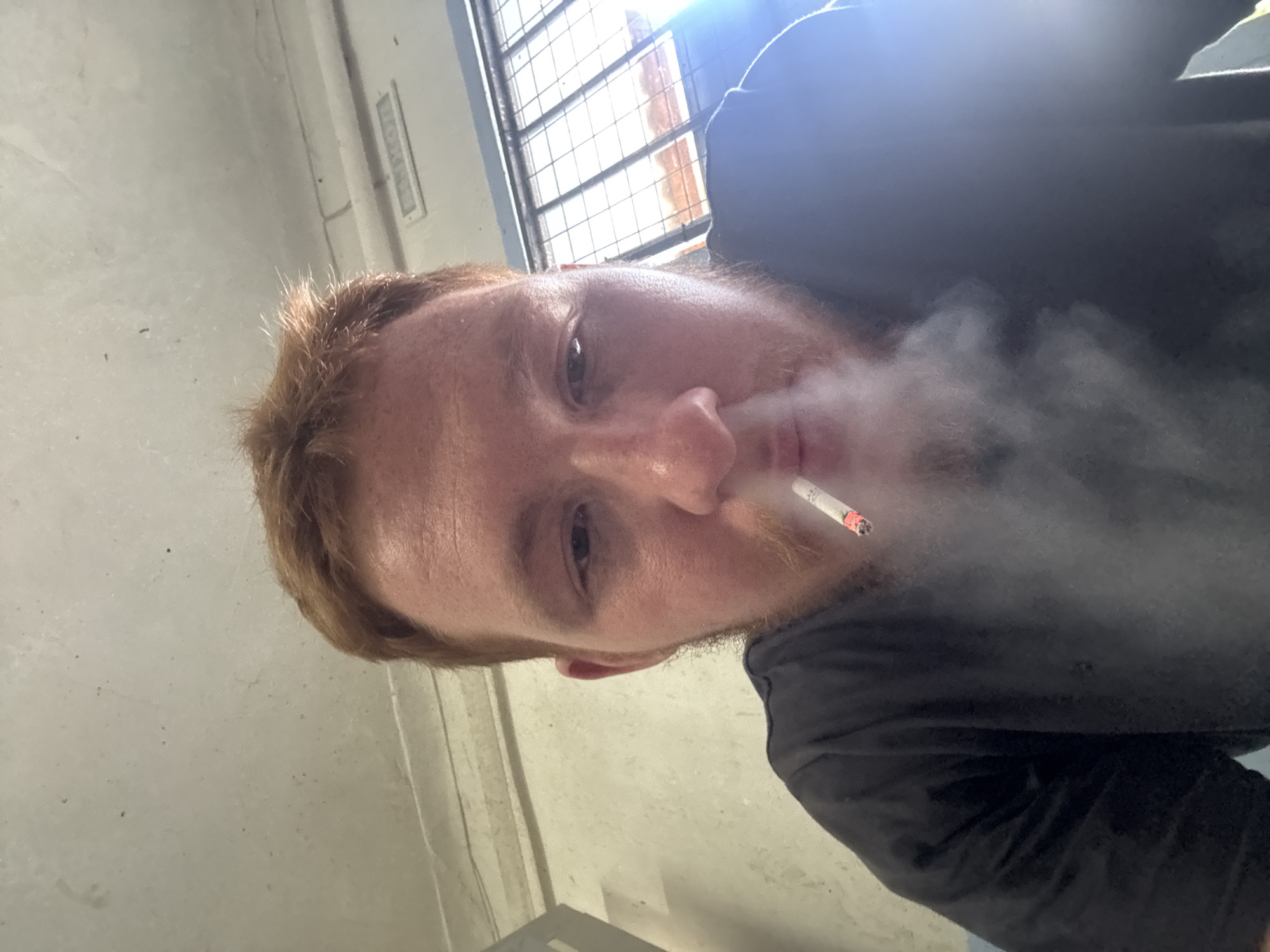 The image is a close-up, selfie-style shot of a person with light-colored skin, reddish-brown hair, and a light beard. The person is smoking a cigarette, with smoke obscuring part of their face and the right side of the image. The subject is wearing a dark t-shirt. The background appears to be an indoor setting with a concrete-like wall on the left and a window with a grid pattern and bright light coming through on the top right. The lighting suggests it could be daytime, as there is strong light from the window. The overall mood is casual, with the smoke creating a somewhat hazy atmosphere. The image is oriented sideways. There is no visible text in the image.