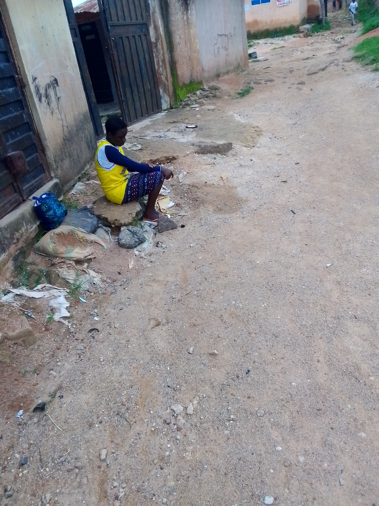 A young person is sitting on a rock next to a building in Jos, Nigeria. The person is wearing a yellow and blue sleeveless shirt and dark patterned shorts. They are looking down with their hands clasped in their lap. To the left of the person is a blue bag and other discarded items. The ground is dirt and gravel. In the background, there is a street with a few other people walking. The building appears to be made of concrete and has a metal door. The overall scene depicts an outdoor urban environment.