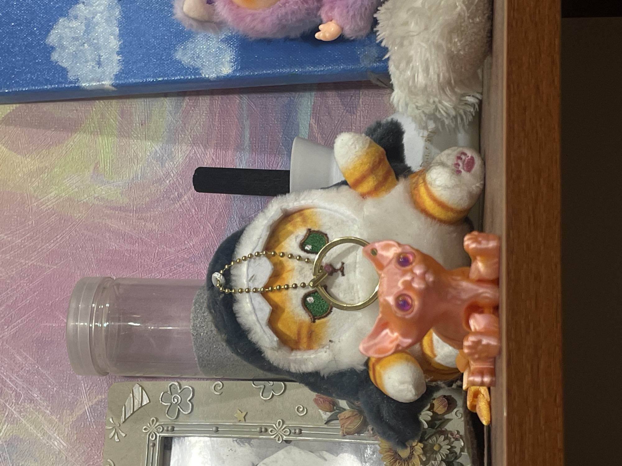 This is a close-up, eye-level shot of a shelf containing various decorative objects. In the foreground, a small, shiny orange figurine of a cat with large, purple eyes sits facing forward. Behind it, a plush cat toy dressed in a grey shark costume with orange stripes is positioned. The cat toy has green eyes and a gold ring hanging from its neck, attached by a delicate gold chain. To the left of the cat toy, a decorative picture frame with floral embellishments is visible. Behind these items, a clear plastic jar filled with small grey pebbles sits next to a black cylindrical object. Further to the right, a portion of a blue canvas painting with white clouds is visible, and to its right, a fuzzy purple toy character with light purple hair and a small hand can be seen. The shelf is made of dark wood, and the background features a textured wall with pink and yellow abstract patterns. The image is set in an indoor environment.