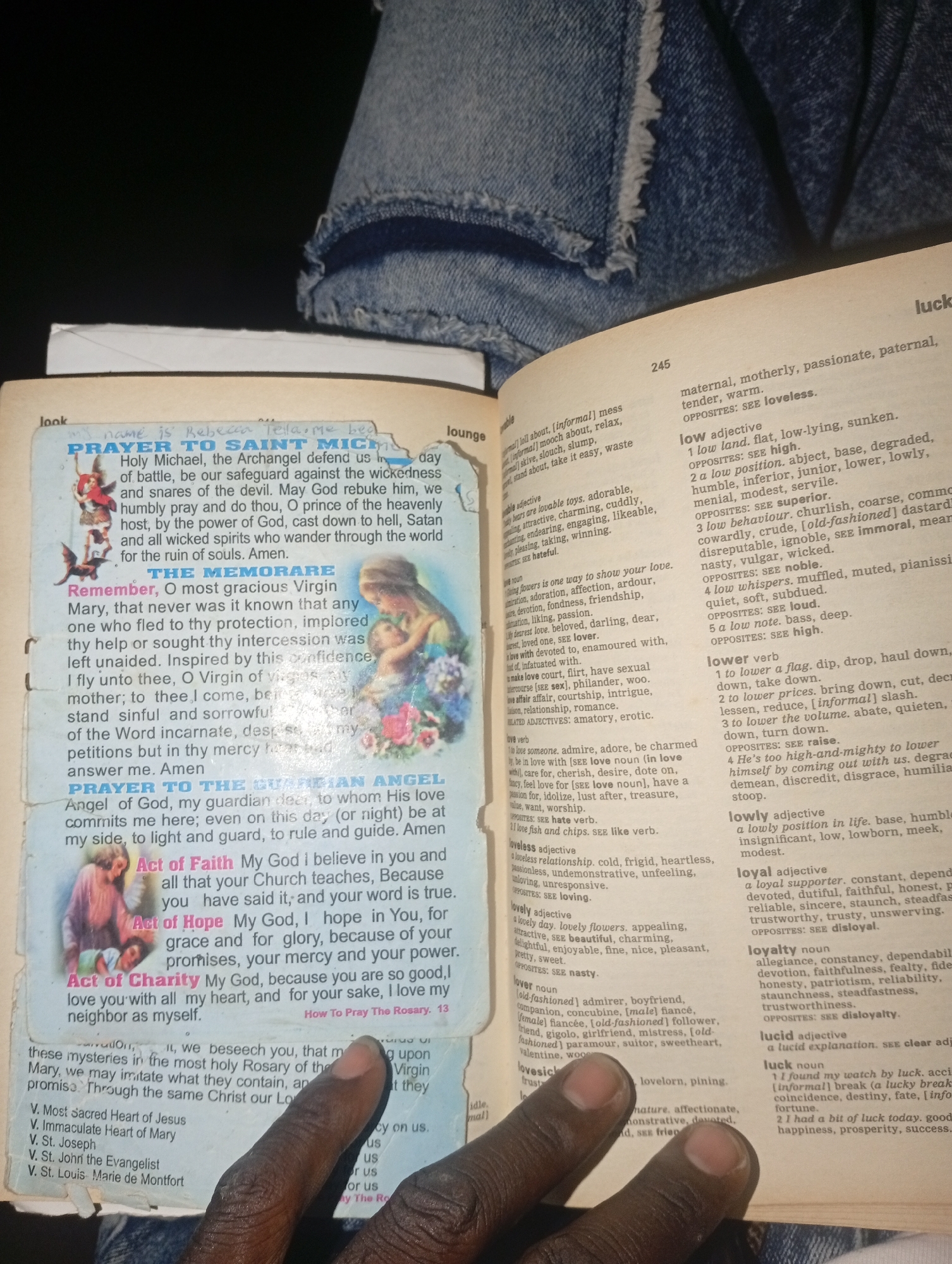 The image shows a book open to pages displaying dictionary entries and religious texts. The book is resting on what appears to be a blue denim surface, likely clothing. On the left page, a prayer to Saint Michael and prayers to the Virgin Mary and Guardian Angel are visible, accompanied by illustrations. The right page contains dictionary definitions for words starting with "l" such as "lovely," "lover," "lovesick," "lucid," and "luck." The text and illustrations are slightly creased and worn. The lighting suggests it is indoors, possibly in a dimly lit environment. There is no discernible information about the city of Gulak, Nigeria, or any specific urban environment.