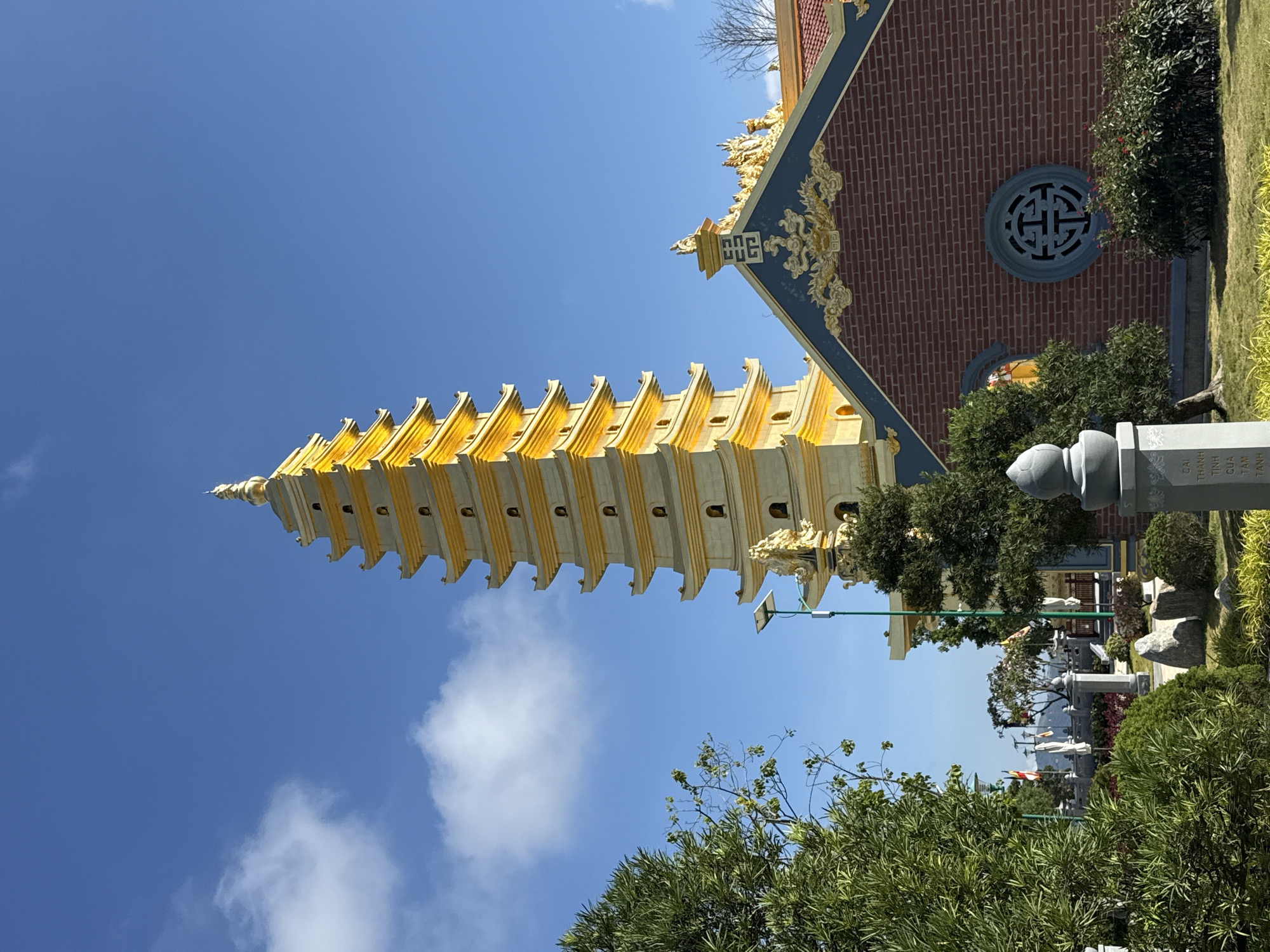 The image shows a multi-tiered, yellow and white pagoda against a clear blue sky with some clouds. The pagoda is situated next to a red brick building with a round decorative window and gold accents. In the foreground, there are trees and vegetation, and a gray concrete structure resembling a memorial or marker. The setting appears to be a temple or park, likely located in Diên Khánh, Vietnam. The photograph is taken from a low angle, emphasizing the height of the pagoda.