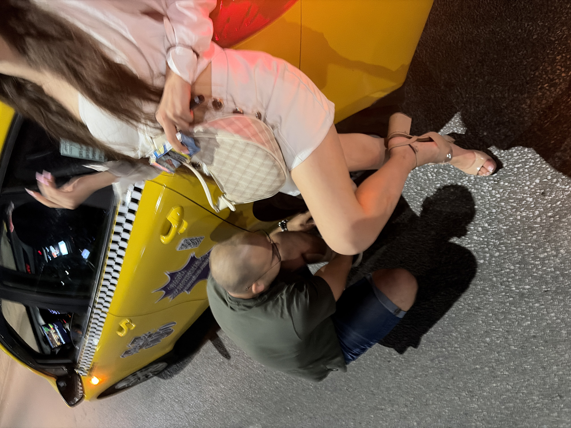 In an outdoor urban environment, a man wearing a dark green t-shirt and denim shorts is crouching down next to a yellow taxi. He is attending to the shoe of a woman who is standing and leaning against the taxi. The woman is wearing a white top and a white skirt, with tan heeled sandals. She is holding a small checkered purse and a small blue package. The taxi has a checkered stripe along its side and graphics on its doors. The ground is asphalt, and shadows are cast from the subjects and the taxi. The lighting suggests it is nighttime.