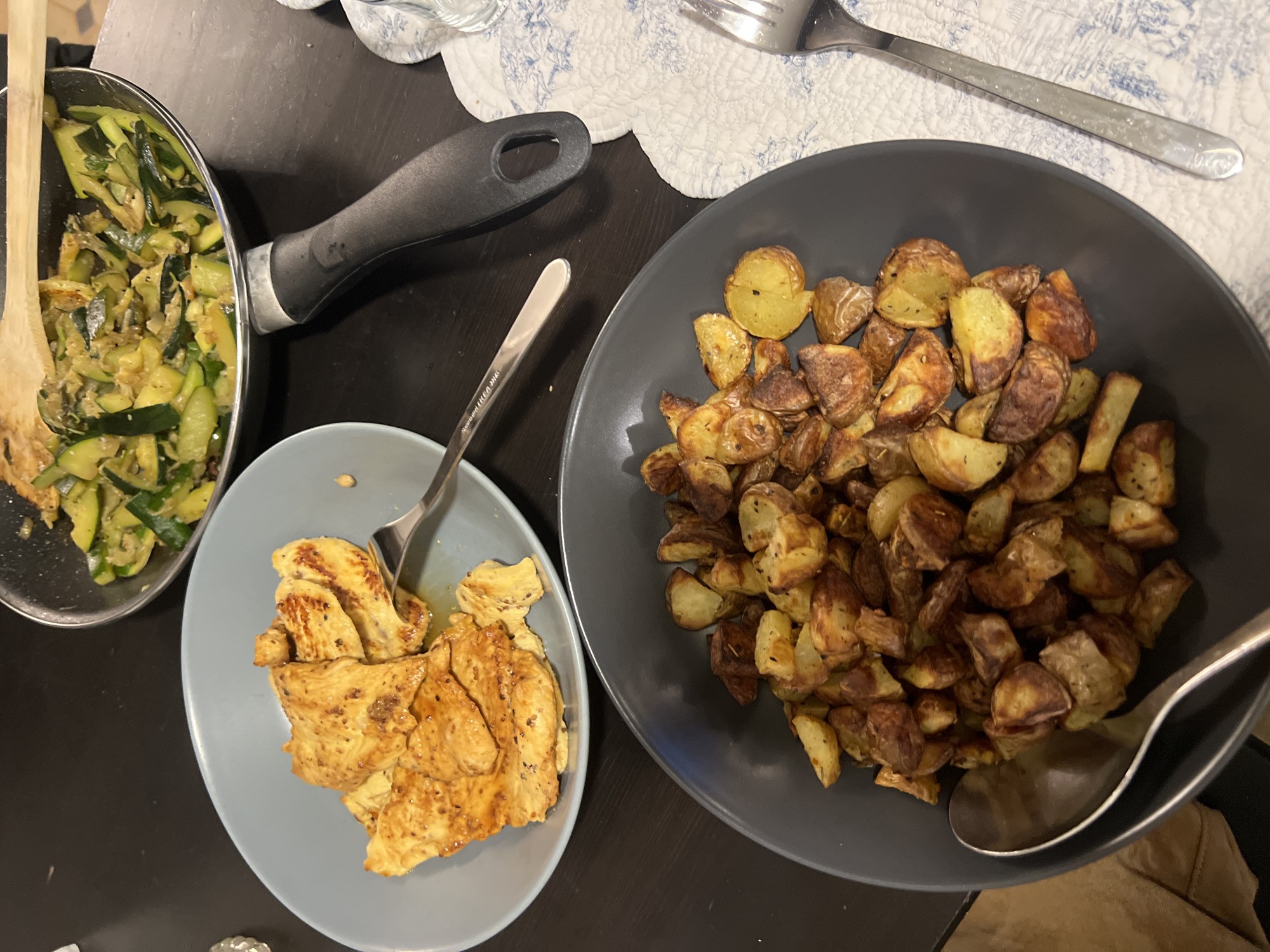 This is a top-down, close-up shot of a meal served on a dark, possibly wooden table. In the foreground, a dark grey bowl is filled with roasted potato wedges. To the left of this bowl, a silver spoon rests inside it, partially obscuring the potatoes. To the right of the bowl, a white placemat with a blue floral pattern is visible, with a silver fork lying on it.
Behind the potato bowl and to the left, a light grey plate holds sliced, pan-fried chicken breast. A fork is also placed on this plate, touching some of the chicken.
In the background, a black frying pan contains sliced zucchini that appears to have been stir-fried. A wooden spatula is resting in the pan, also touching the zucchini. The scene depicts a home-cooked meal. There is no information in the image to indicate the location in Paris, France.