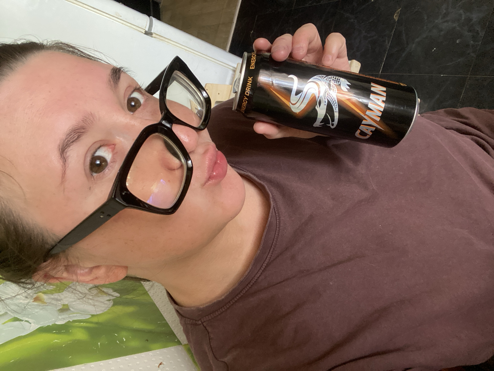 A woman with dark hair, wearing large black-rimmed glasses and a brown t-shirt, holds a black can of "CAYMAN ENERGY DRINK" with a silver alligator design. She is looking into the camera, puckering her lips. The background includes a white refrigerator, a patterned wall covering with green and white floral motifs, and a dark tiled floor. The image is taken from a slightly high angle, looking down at the subject. The overall setting appears to be an indoor domestic environment. There is no discernible information in the image that places the scene in the city of Chaikovsky, Russia.