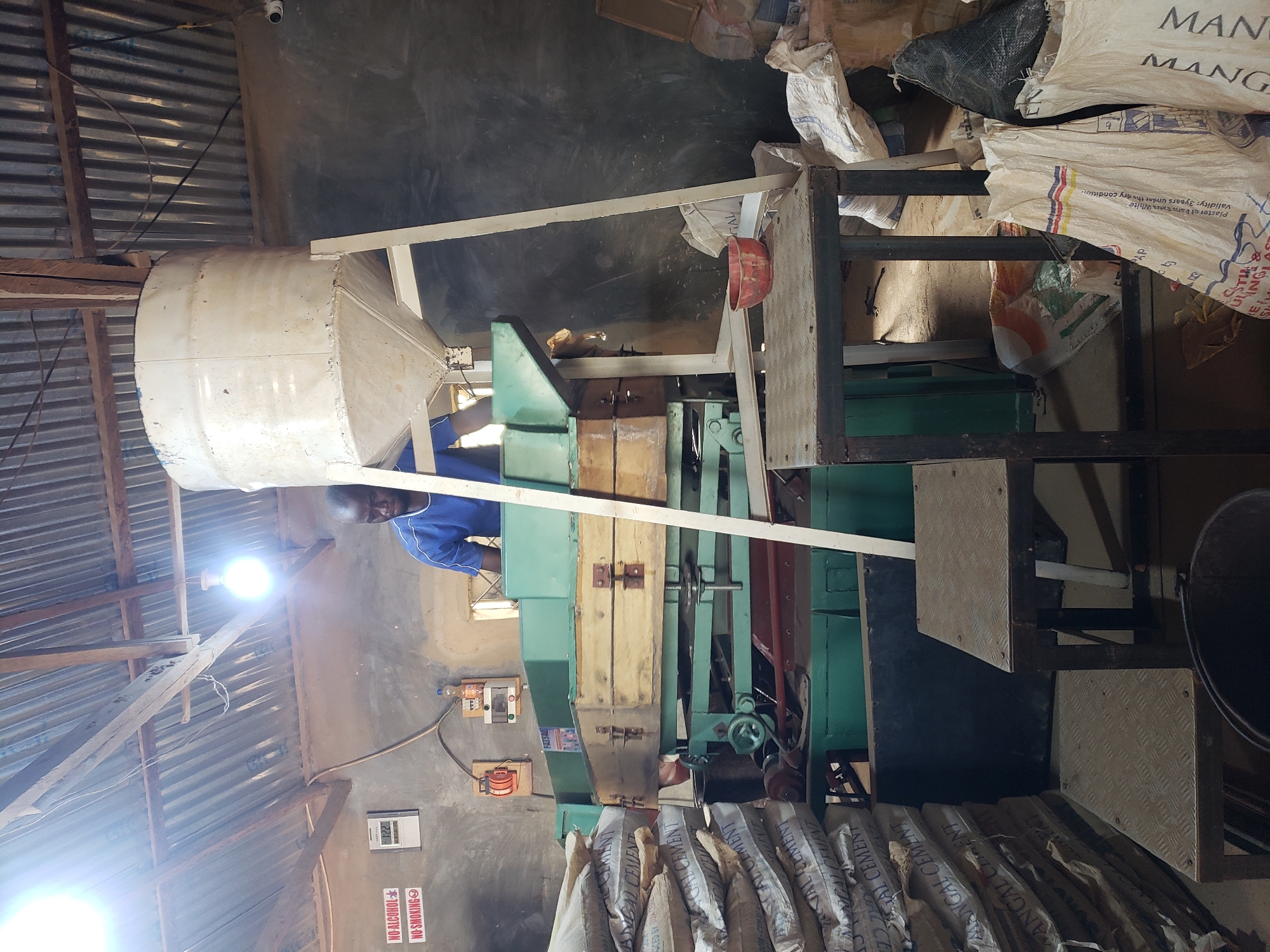The image depicts an interior scene, likely within a workshop or processing facility in Jos, Nigeria. A man, wearing a blue t-shirt, is standing behind a large piece of industrial machinery, which appears to be a sifting or processing unit. This machine is green and has wooden components, with various knobs and levers suggesting operational controls. Above the machinery, a tall, white conical hopper is supported by a metal frame.
To the left of the man and machine, stacks of large sacks are piled high. These sacks are light-colored and appear to be filled with a granular substance, with markings such as "CEMENT" visible on some. A digital clock displaying "12:22" and two signs, one with "NO ALCOHOL" and another with "NO SMOKING" and a circled "X", are mounted on the wall behind the machinery.
In the foreground, a metal stand with two diamond-plate steps is positioned. On the top step rests a small red bowl. To the right of this stand, more sacks are visible, some appearing to be made of rough material and others of a lighter woven fabric, with text and graphics on them. The overall environment suggests an active production or storage area, with industrial equipment and bagged materials. The ceiling is corrugated metal, and the walls are a neutral, muted color.