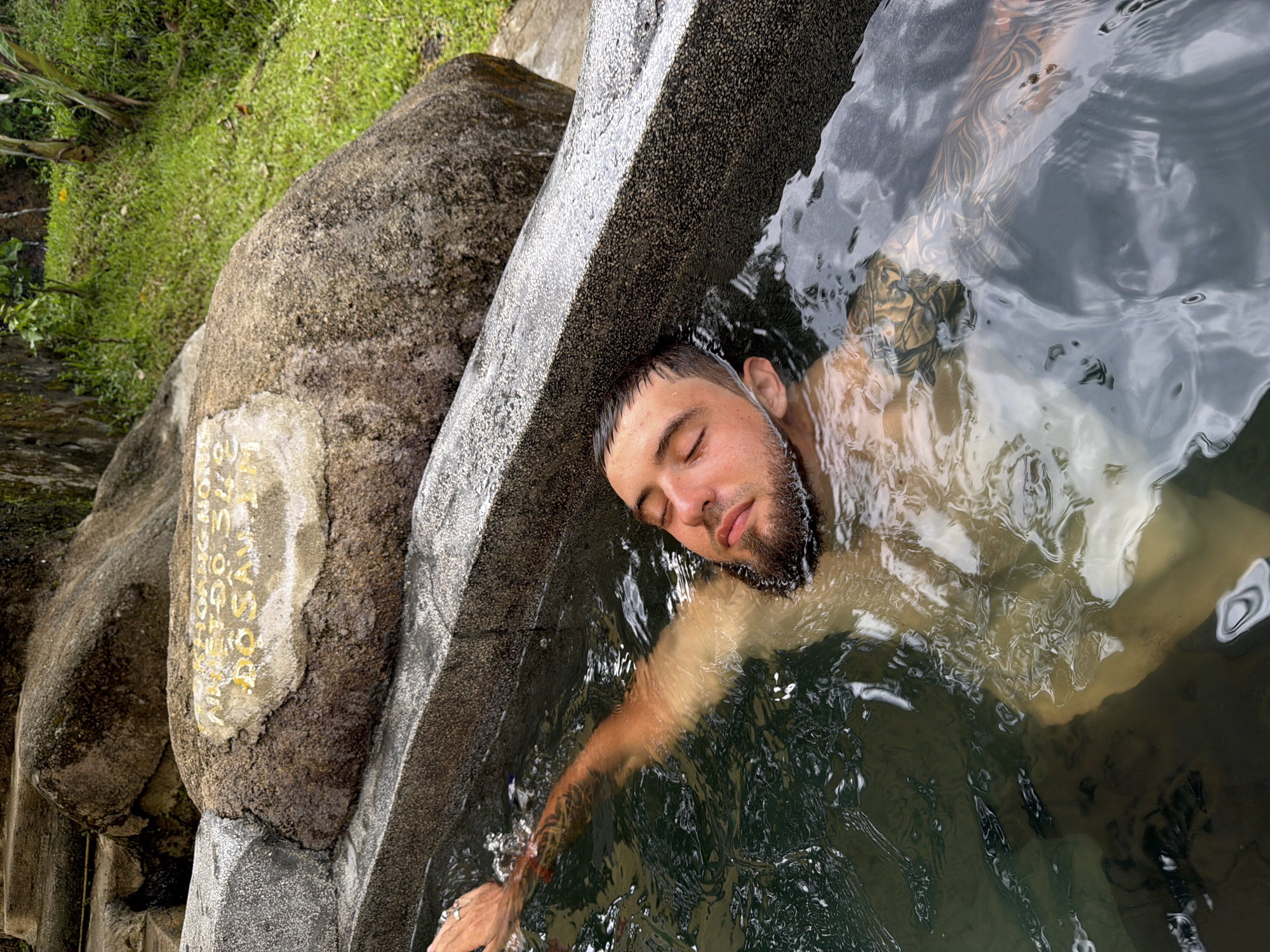A man with dark hair and a beard is partially submerged in a stone hot spring, his eyes closed and his right arm extended above the water. He has tattoos on his arm and chest that are visible through the water. The hot spring is surrounded by large rocks and lush green vegetation. In the background, there are stone steps leading up a grassy slope. A weathered stone plaque is embedded in a large rock near the edge of the hot spring. The plaque appears to have yellow lettering, but the text is difficult to decipher. The setting is likely an outdoor natural area in or around Khanh Vinh, Vietnam, known for its hot springs.