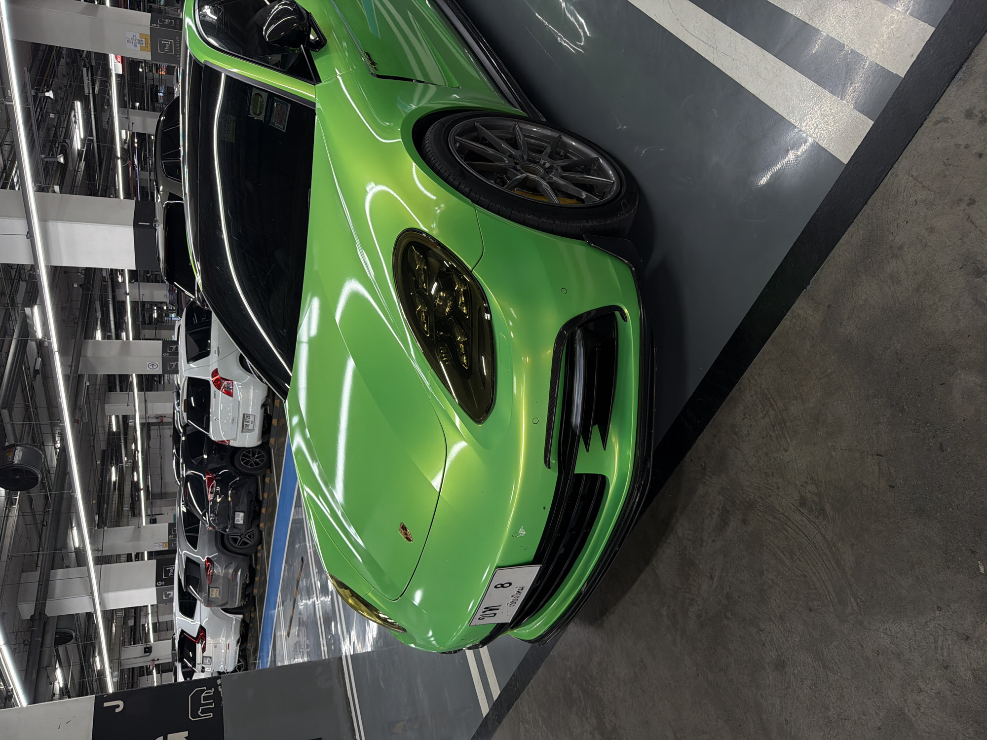 In a well-lit, multi-level parking garage in Nong Pa Khrang, Thailand, a vibrant, metallic green Porsche Panamera is parked. The car is positioned at an angle, showcasing its front and right side. The license plate, which appears to be Thai, is visible on the front bumper. Other vehicles, including white SUVs, a black SUV, and a white hatchback, are parked in the background. The floor of the garage is a mix of concrete and painted lines, indicating parking spaces. The ceiling features fluorescent lighting and exposed infrastructure. The overall environment is clean and organized. There are no people visible in the image.